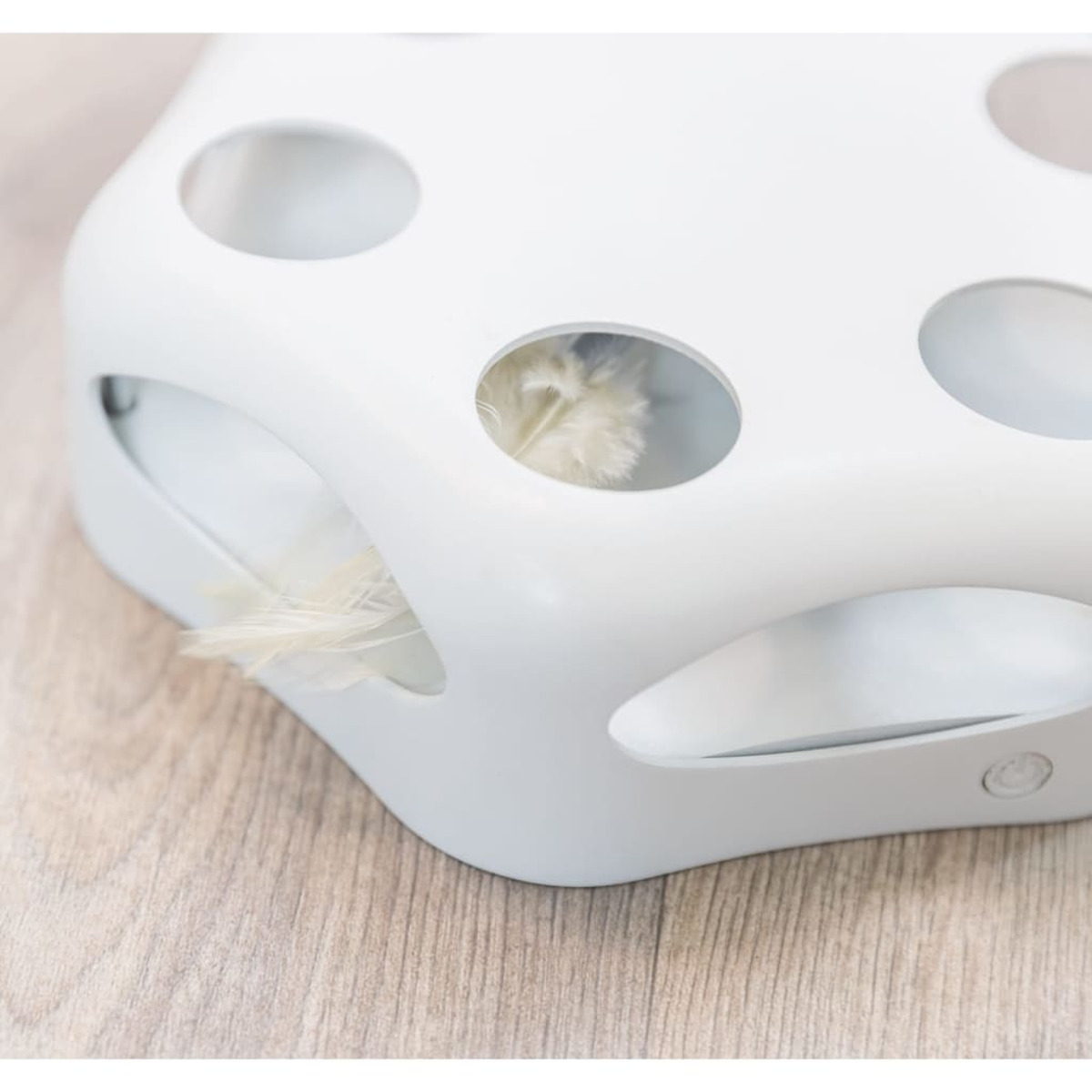 Biała zabawka dla kota z otworami i piórem w środku. Na drewnianej powierzchni.