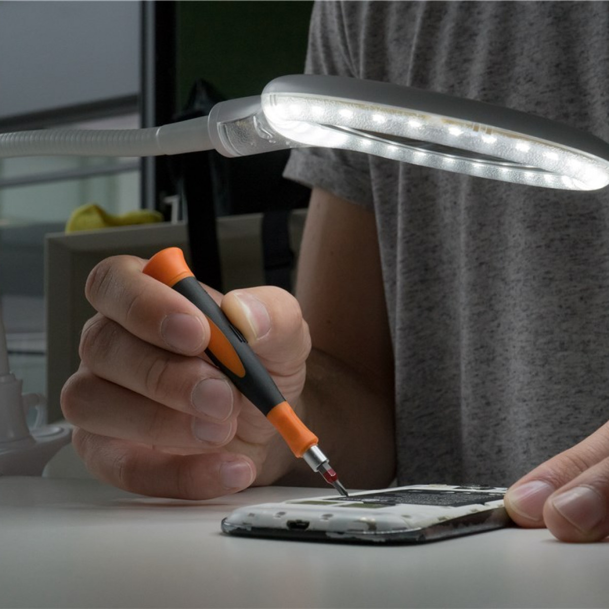 Person repariert ein Telefon mit Schraubendreher und heller Schreibtischlampe.