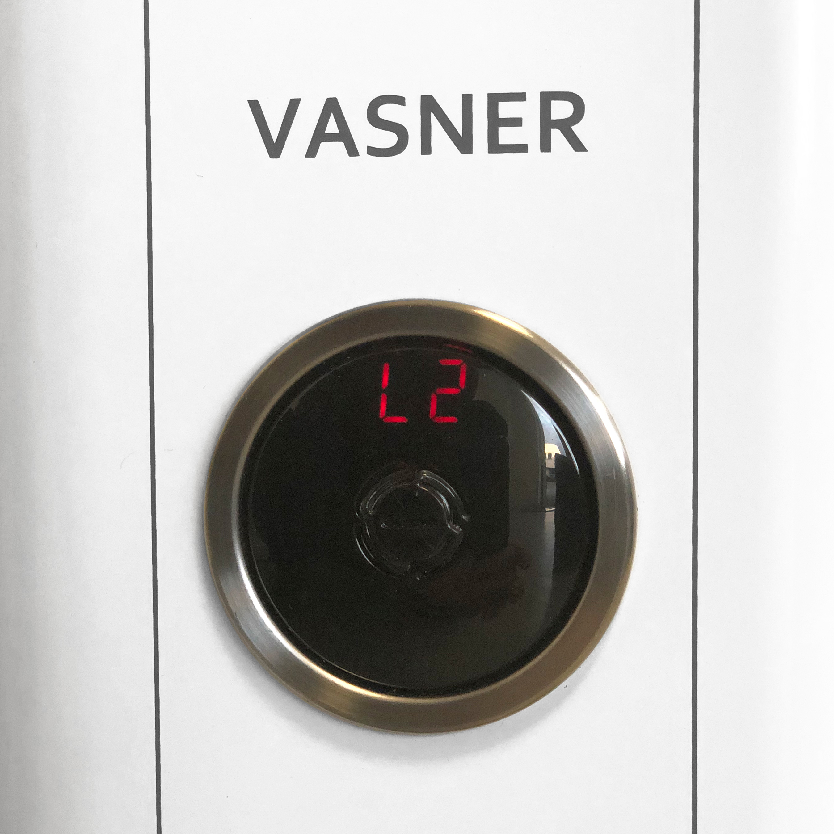 Eine weiße Oberfläche mit einem schwarzen, runden Zifferblatt mit einer Digitalanzeige mit 'L2'. Die Marke ist VASNER.
