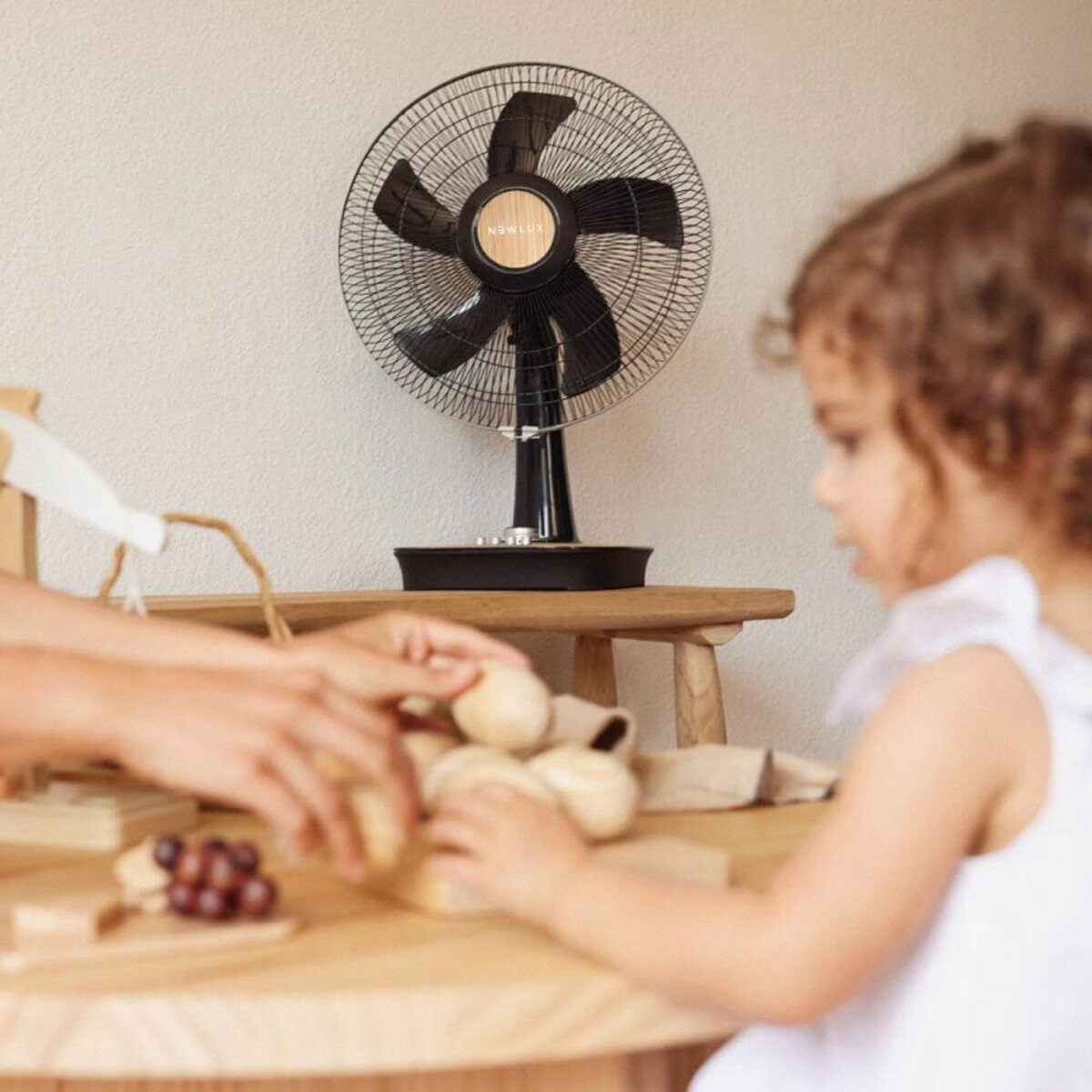 Un niño y un adulto juegan con juguetes de madera frente a un ventilador negro sobre una mesa. Pared blanca al fondo.
