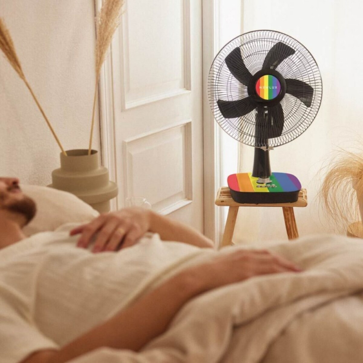 Una persona acostada en la cama con un ventilador con diseño arcoíris en un taburete.