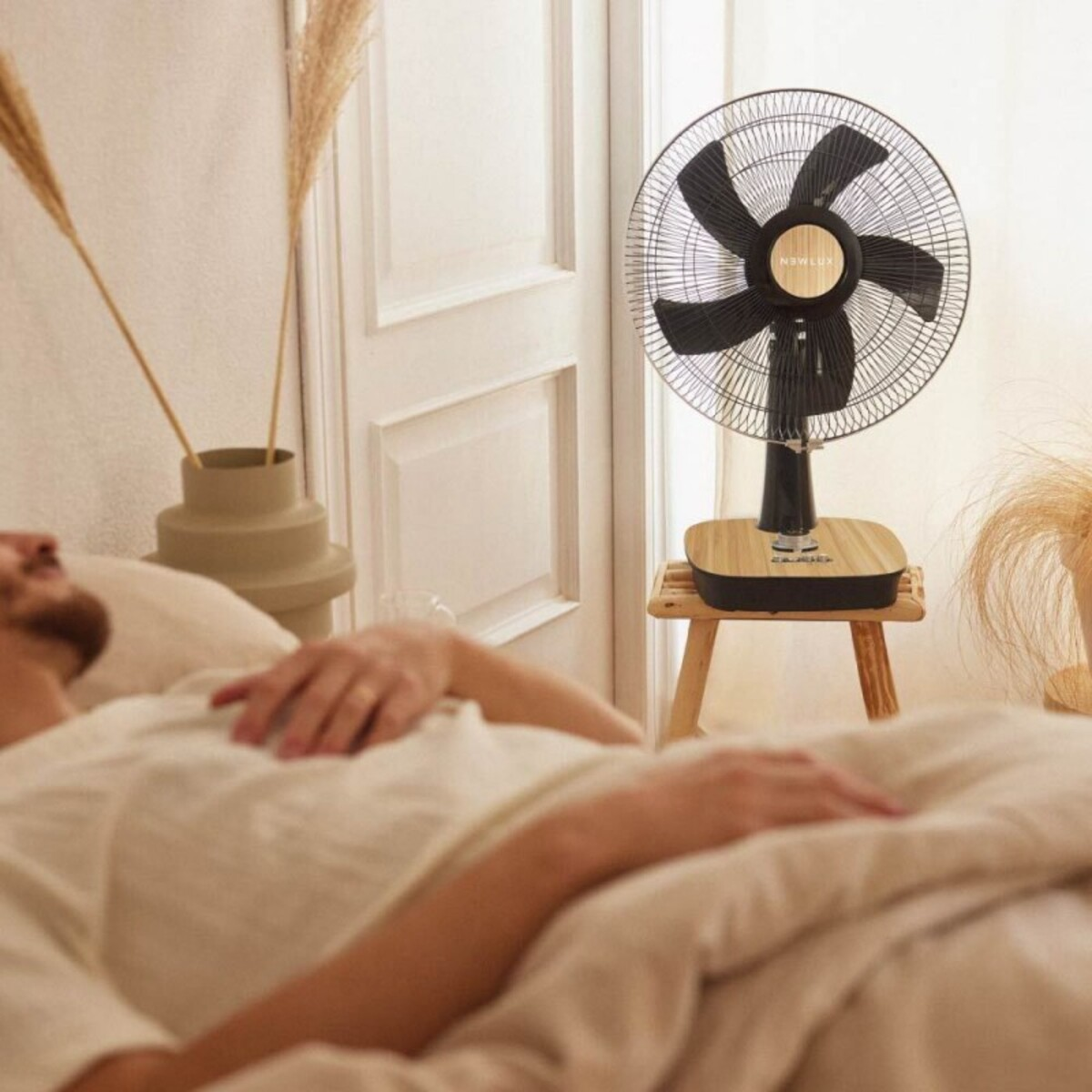 Un hombre en la cama y un ventilador en una mesita. Un jarrón con plantas a la izquierda.