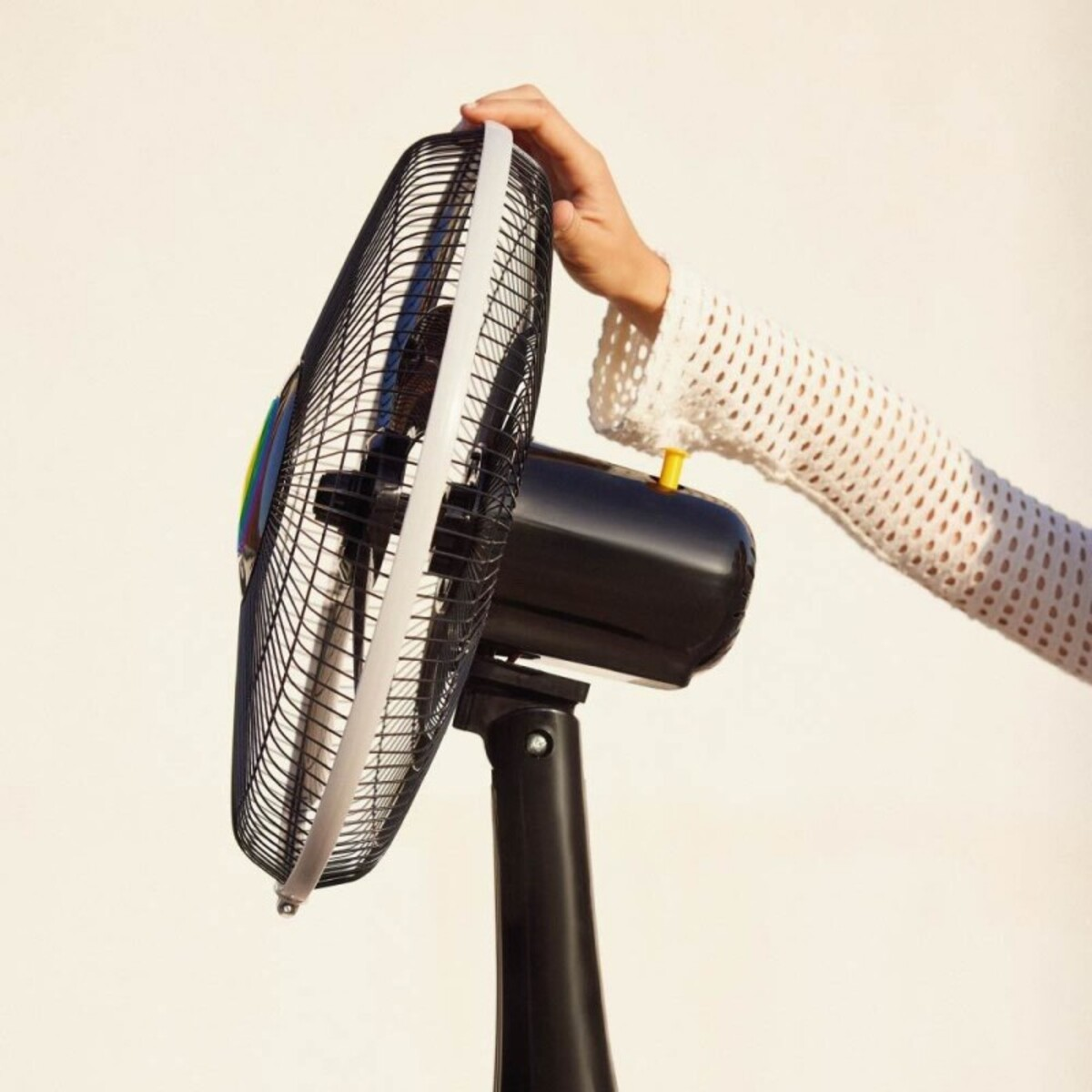 La mano de una persona ajustando un ventilador eléctrico negro con borde blanco, sobre fondo blanco.