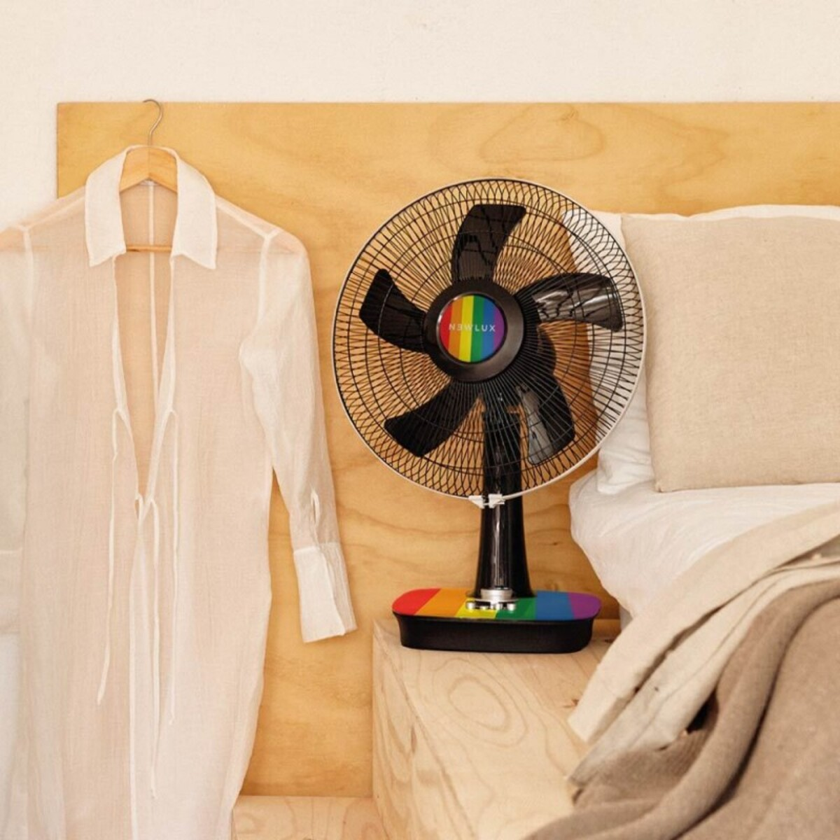 Un ventilador con un logotipo de arcoíris en una mesita de noche junto a una cama con una camisa blanca colgando.