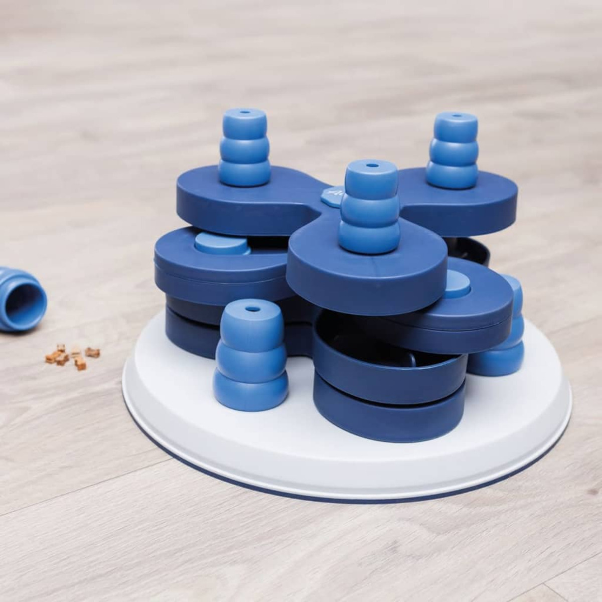 Giocattolo puzzle blu per cani con più livelli e pioli su base bianca, pavimento in legno.