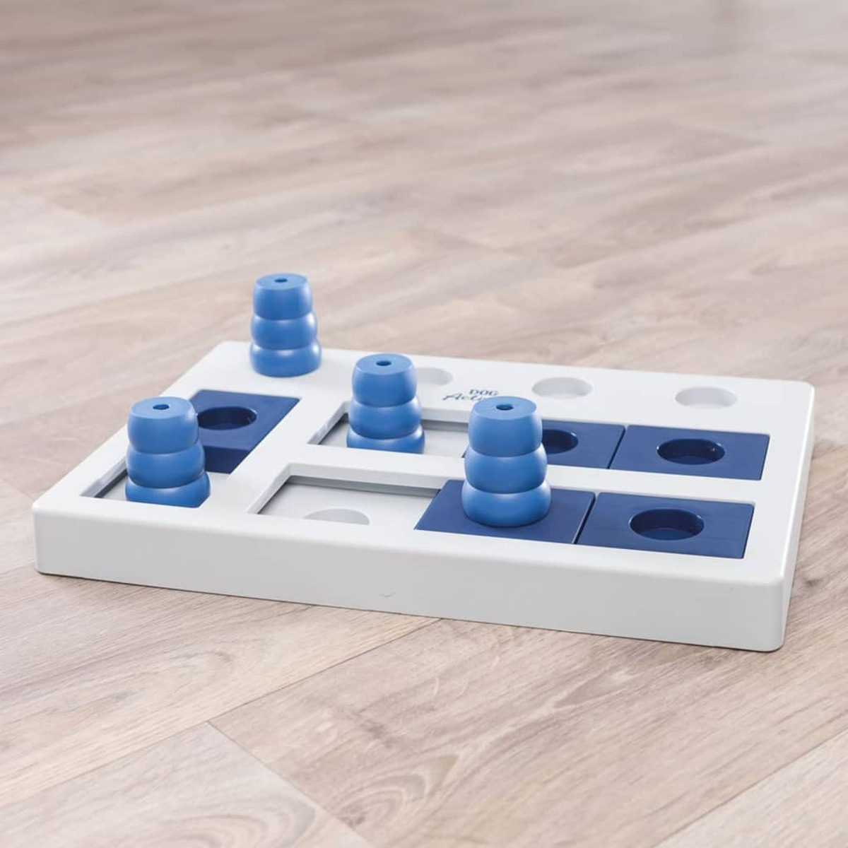 Giocattolo puzzle per cani. Base in plastica bianca e blu con torri blu e fori quadrati. Pavimento in legno chiaro.