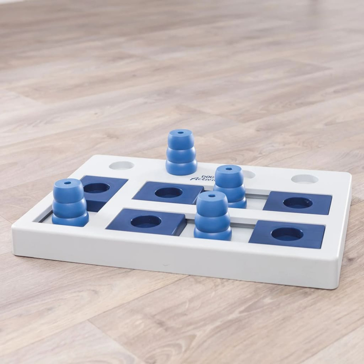 Un giocattolo puzzle per cani con pioli blu e una base bianca. Il giocattolo è su un pavimento di legno.