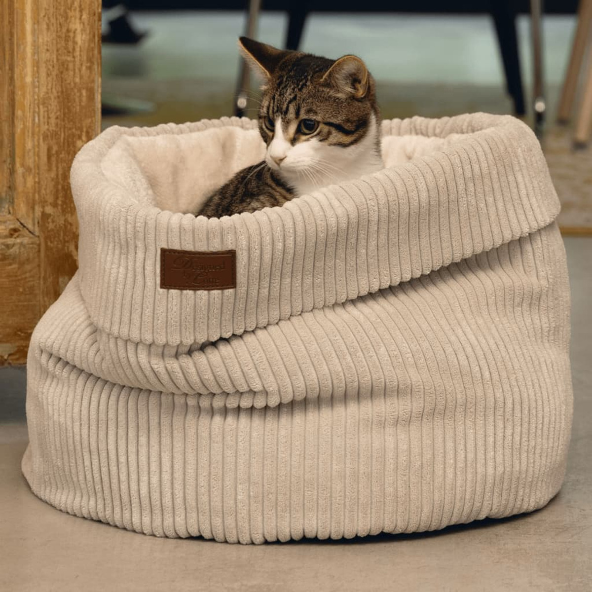 Eine getigerte Katze sitzt in einem Cord-Haustierbett. Das Bett ist hellbeige und hat ein braunes Etikett.