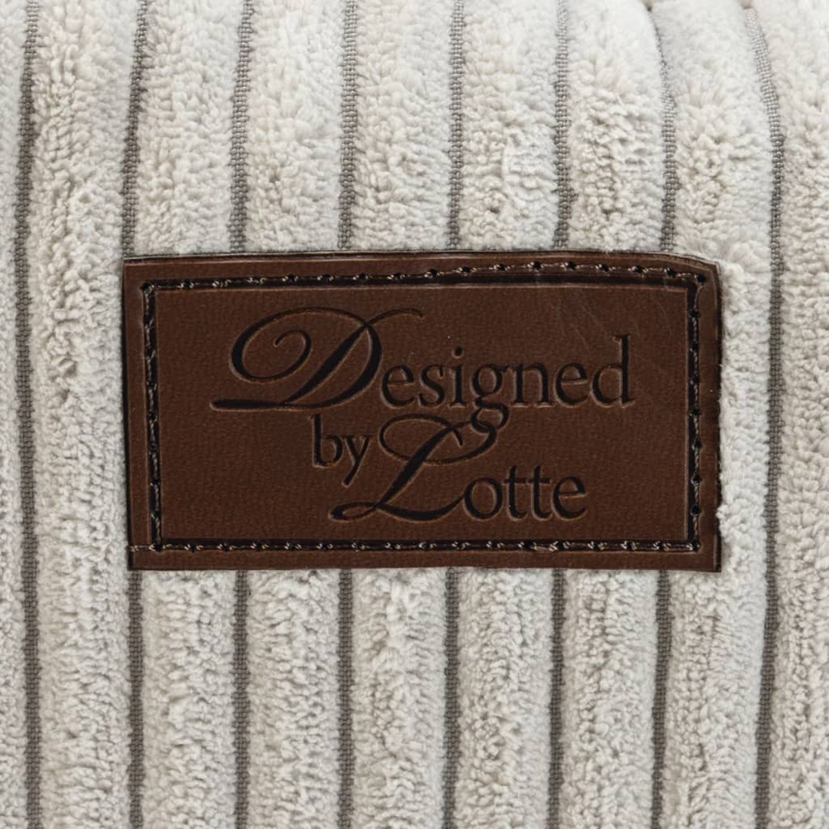 Ein braunes Lederetikett auf einem strukturierten, gestreiften Stoff. Das Etikett lautet "Designed by Lotte".