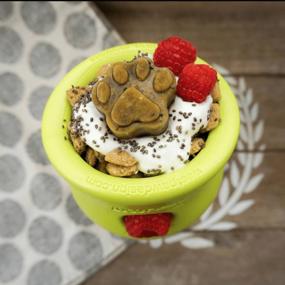 Una ciotola verde per cani con un dolcetto a forma di zampa sopra lo yogurt e circondato da crocchette e lamponi.