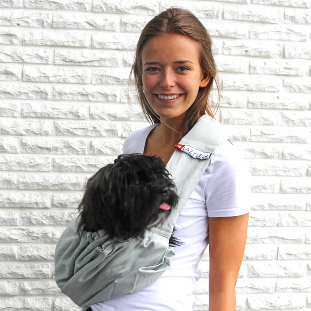 Une femme avec un petit chien noir dans un sac en bandoulière gris clair. Le chien tire la langue.