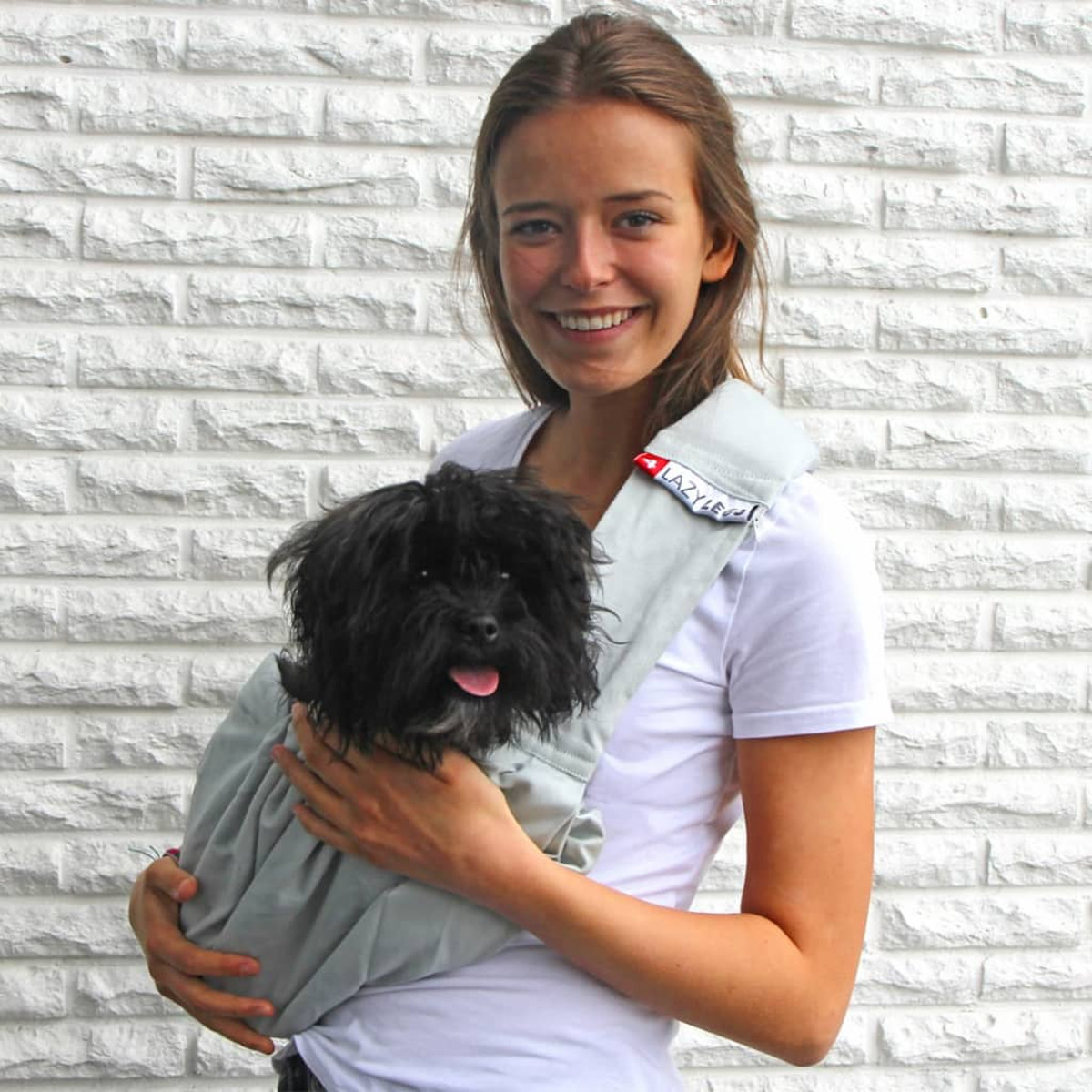 Une femme souriante tenant un petit chien noir dans un sac de transport gris, contre un mur de briques.