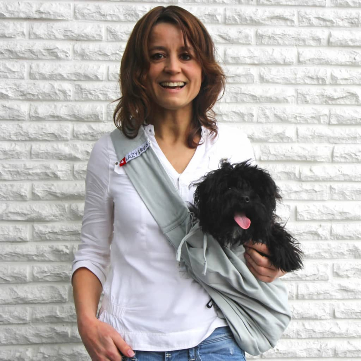 Eine Frau mit Hund in einer grauen Trage, lächelt in die Kamera, weißer Ziegelhintergrund.
