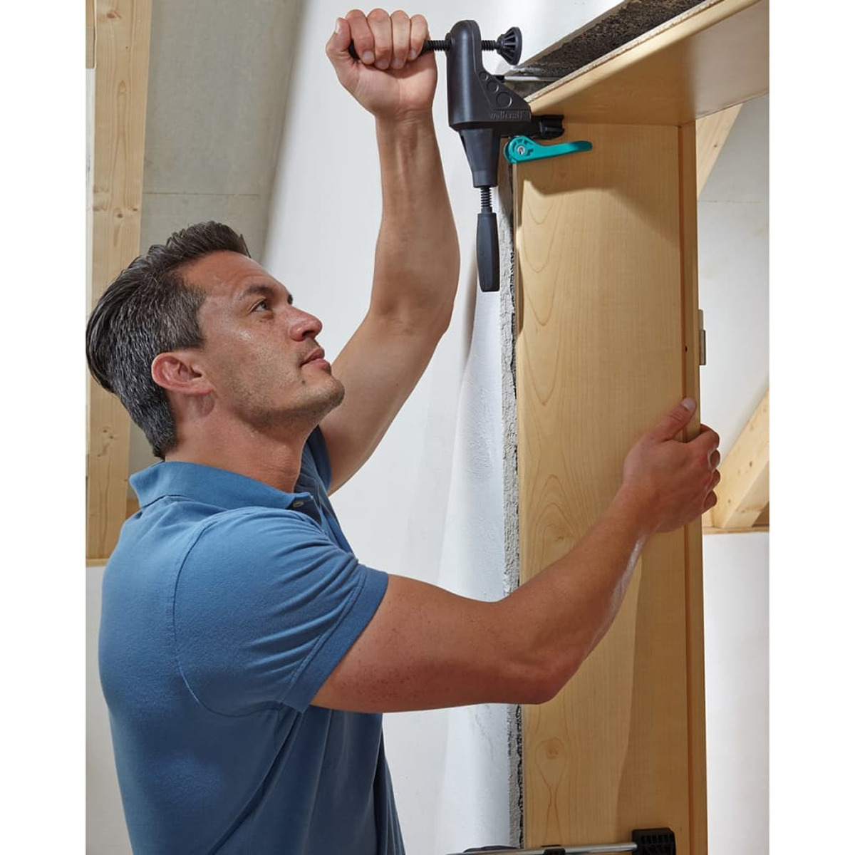 Ein Mann befestigt mit einer Klemme eine Holzplatte an einer Wand. Er trägt ein blaues Hemd.