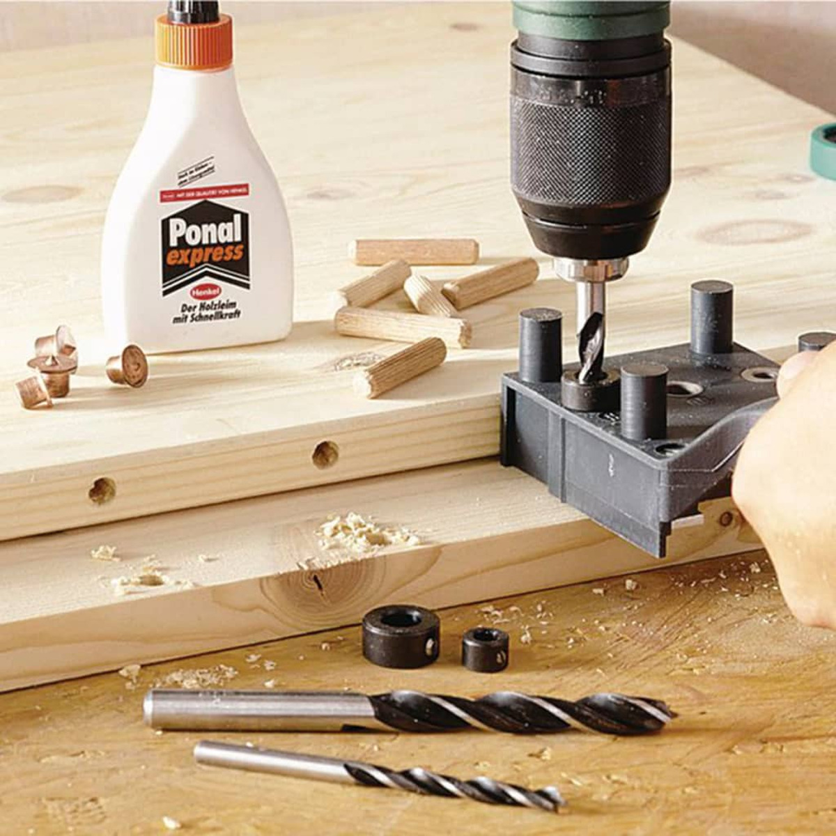 Scène de menuiserie : colle, perceuse, chevilles et mèches sur bois. Les outils sont en usage, avec des copeaux.