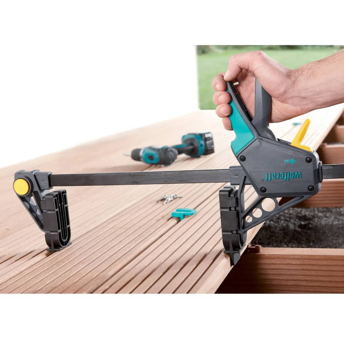 La mano di un uomo usa un morsetto per fissare assi di legno. Un trapano e altri attrezzi sono visibili sullo sfondo.
