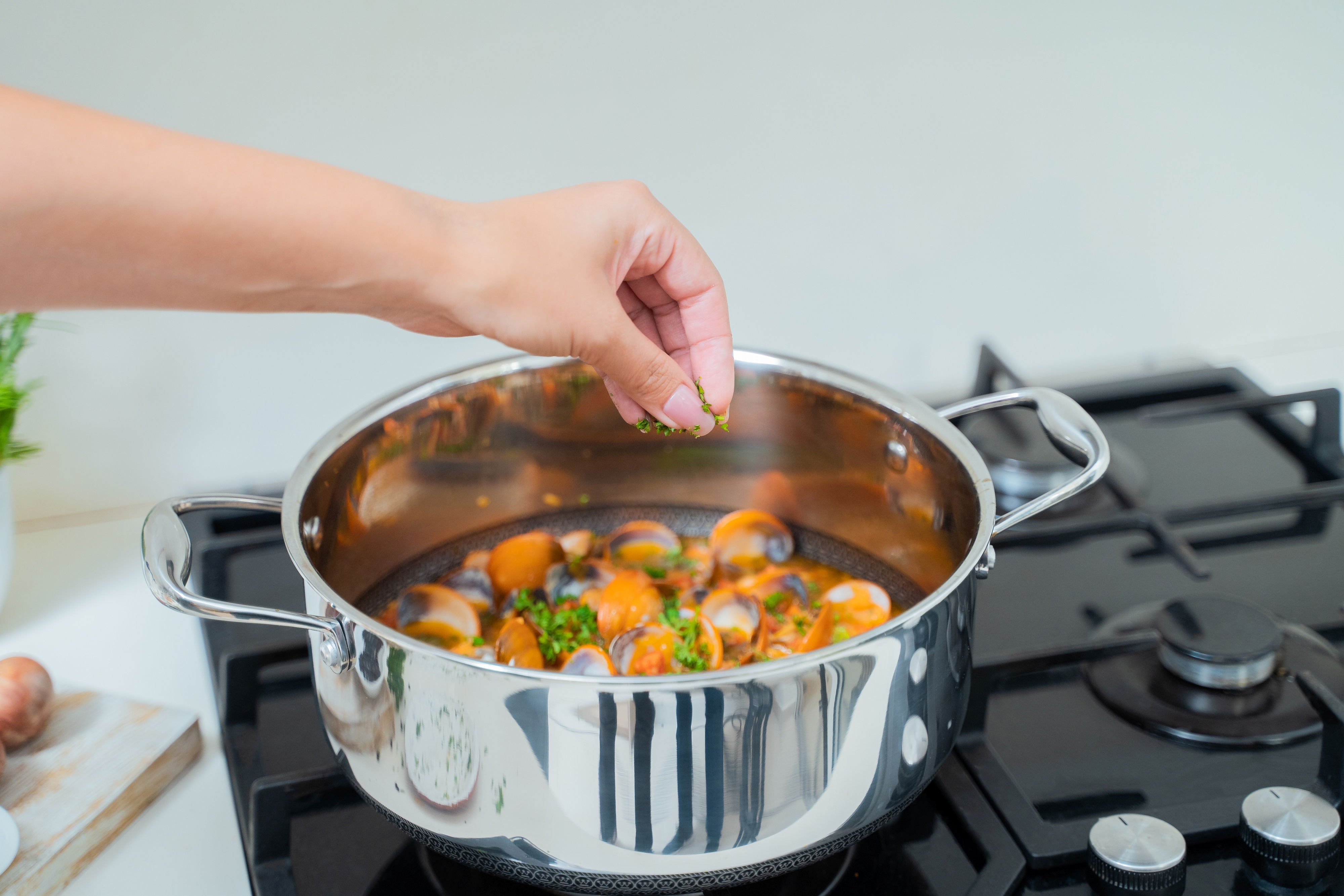 Eine Person gibt Kräuter zu einem Topf mit Muscheln in Tomatensauce auf einem schwarzen Herd.