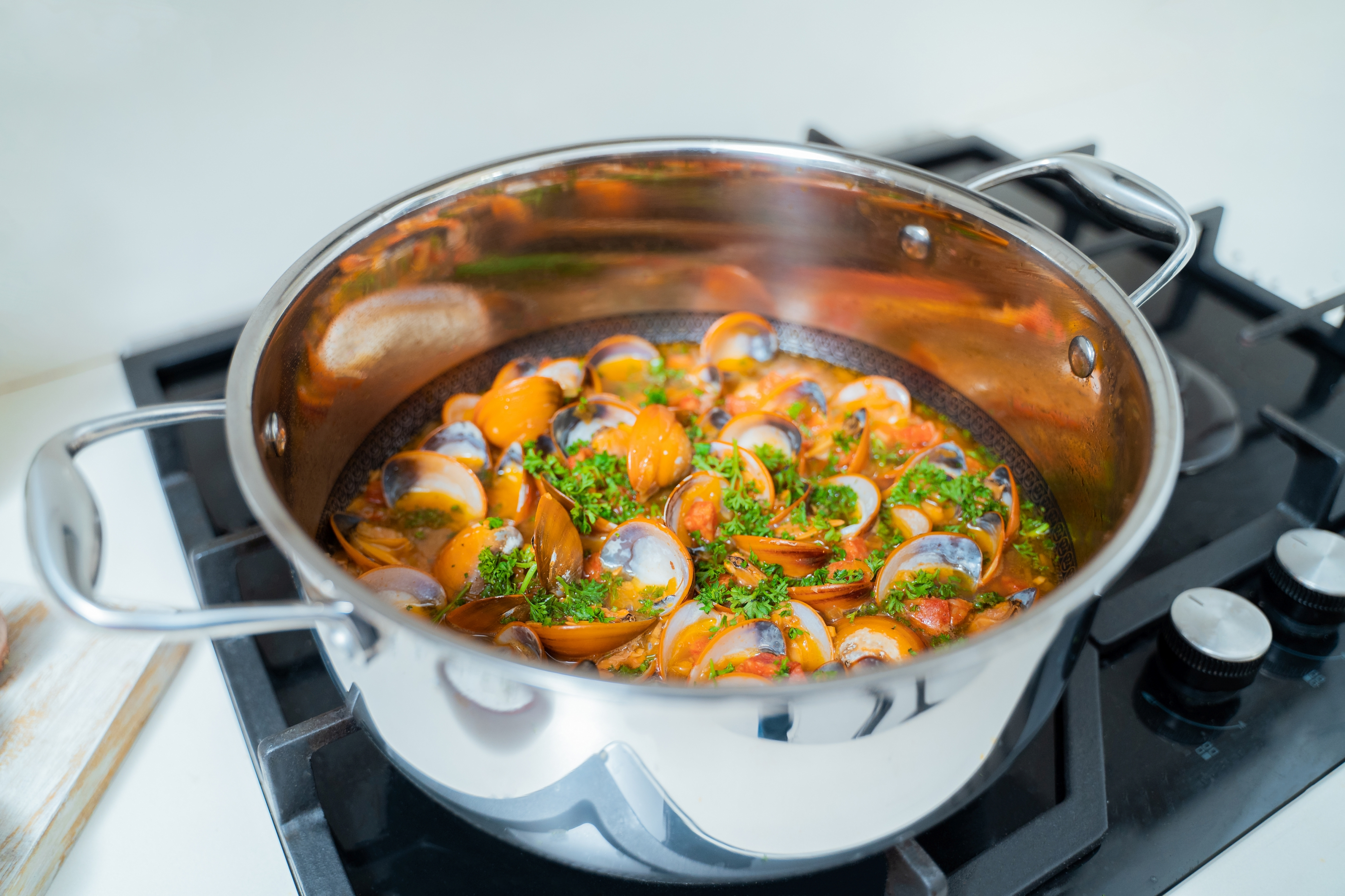 Muscheln in einem Edelstahltopf auf einem schwarzen Herd. Grüne Kräuter obendrauf. Kochen in Bearbeitung.