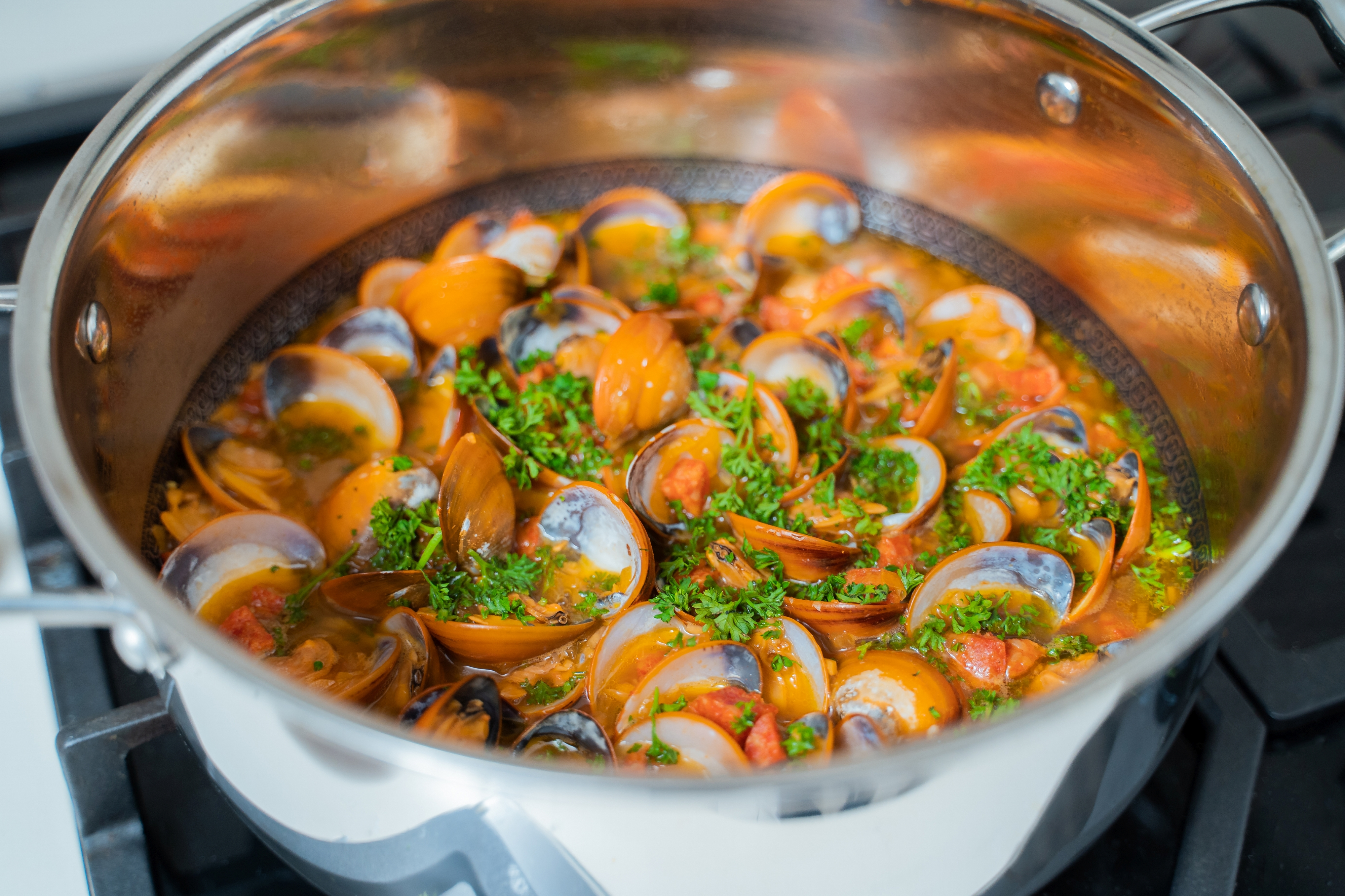Ein Topf Muscheln mit Tomatensauce und Kräutern. Die Muscheln sind geöffnet und orange, mit grünen Kräutern obenauf.
