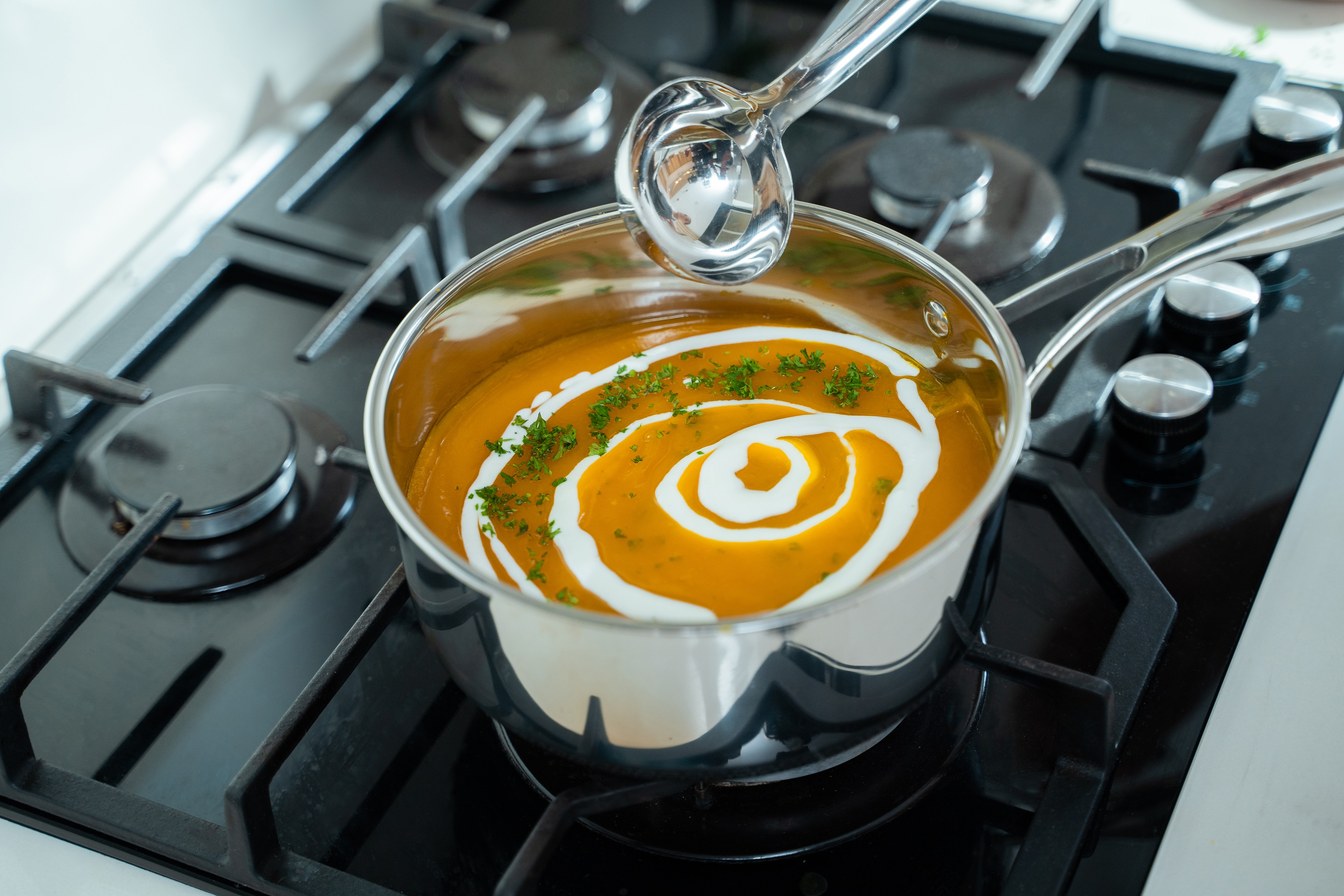 Ein Topf mit orangener Suppe mit weißen Sahnewirbeln auf einem schwarzen Herd. Eine Kelle darüber.