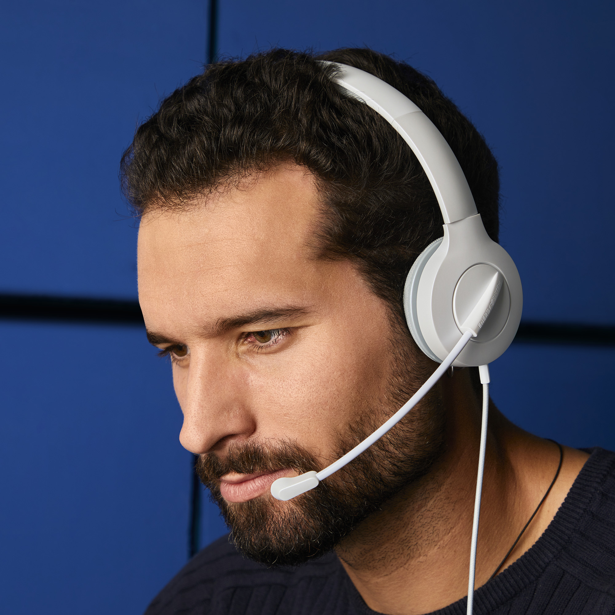 Hombre con auriculares blancos y micrófono, mirando algo, sobre fondo azul.