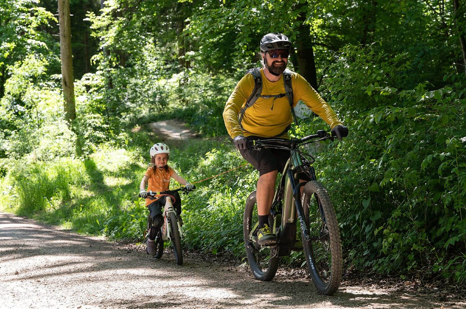 Vater und Kind fahren mit dem Fahrrad auf einem Waldweg. Das Fahrrad des Kindes ist mit dem des Vaters verbunden.