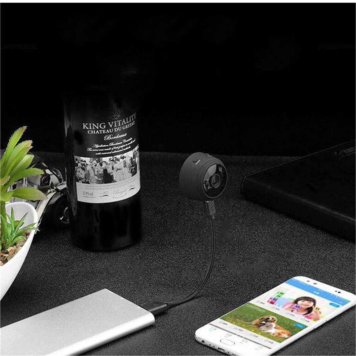 Mini cámara espía, botella de vino, teléfono mostrando video y un banco de energía en una superficie oscura.