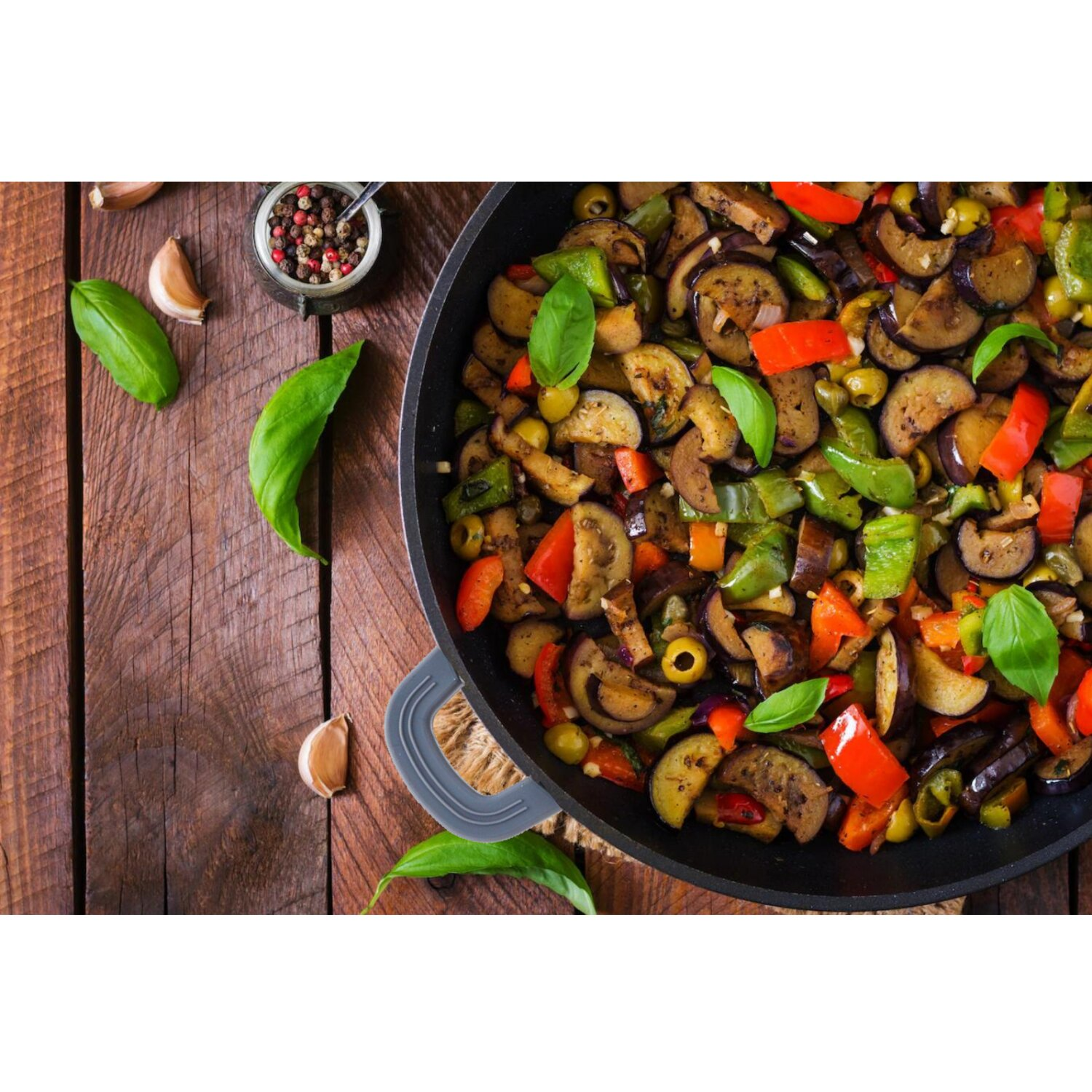 Draufsicht auf Ratatouille in einer schwarzen Pfanne. Drumherum Basilikumblätter, Knoblauchzehen und Pfefferkörner.