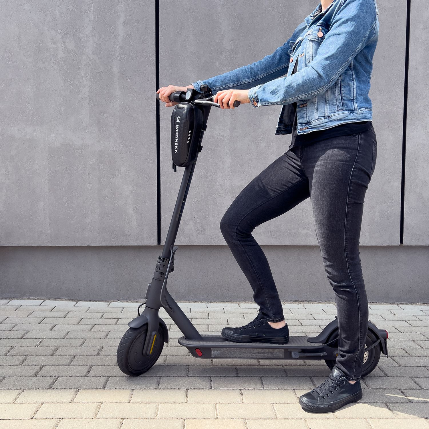 Eine Person auf einem Roller, trägt eine Jeansjacke und schwarze Hose. Graue Wand.