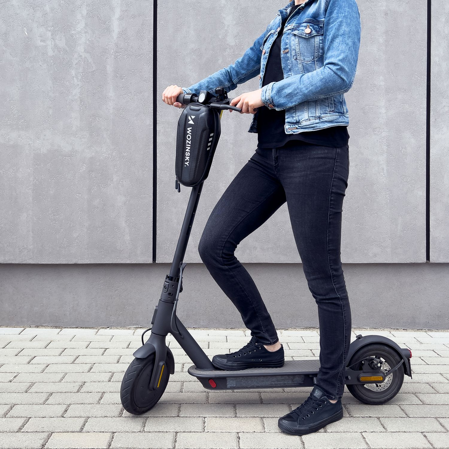 Eine Person fährt einen Elektroroller auf einer Ziegeloberfläche. Sie tragen Schwarz und Jeans.