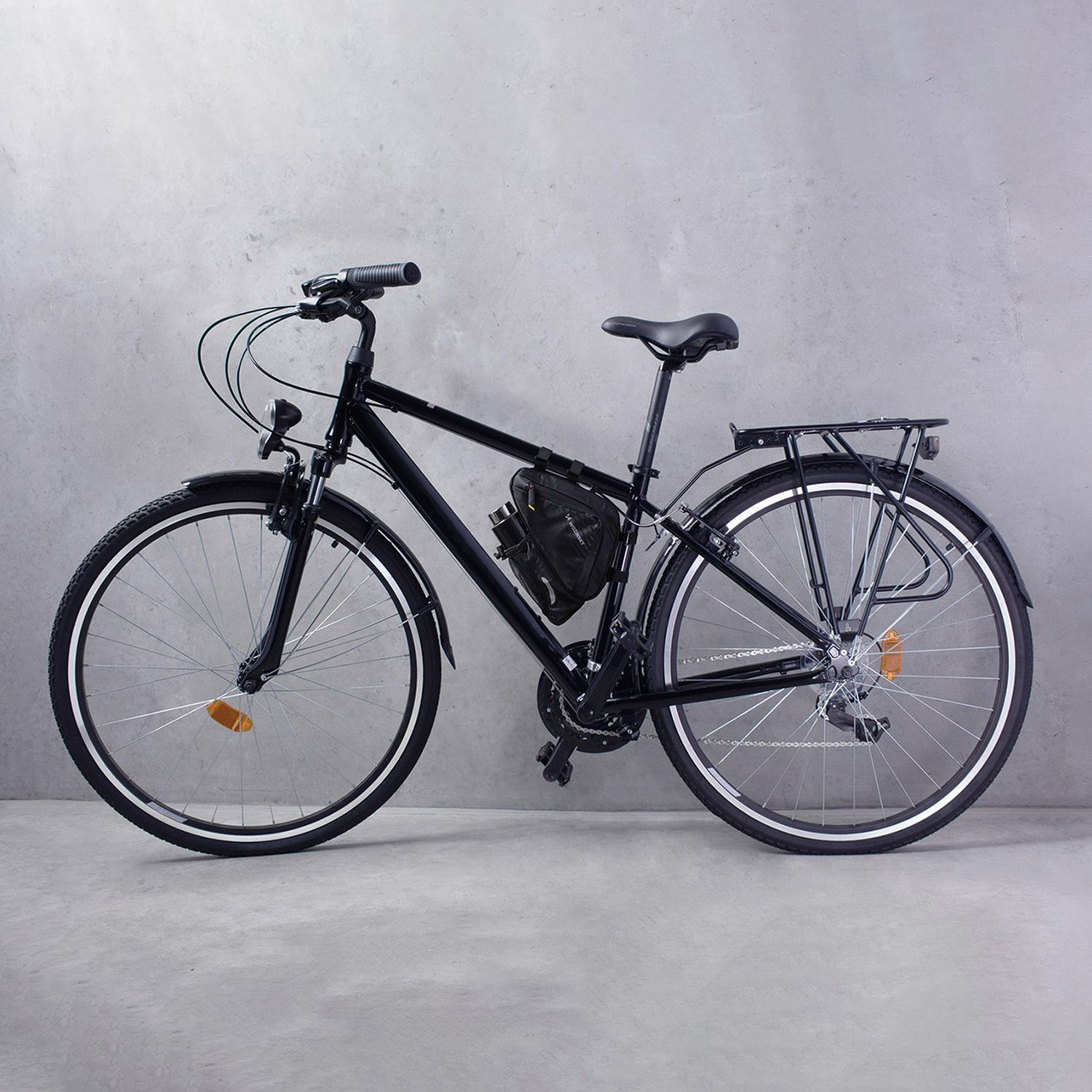 Vélo noir avec jantes grises et sac noir sur le cadre. Appuyé contre un mur gris.