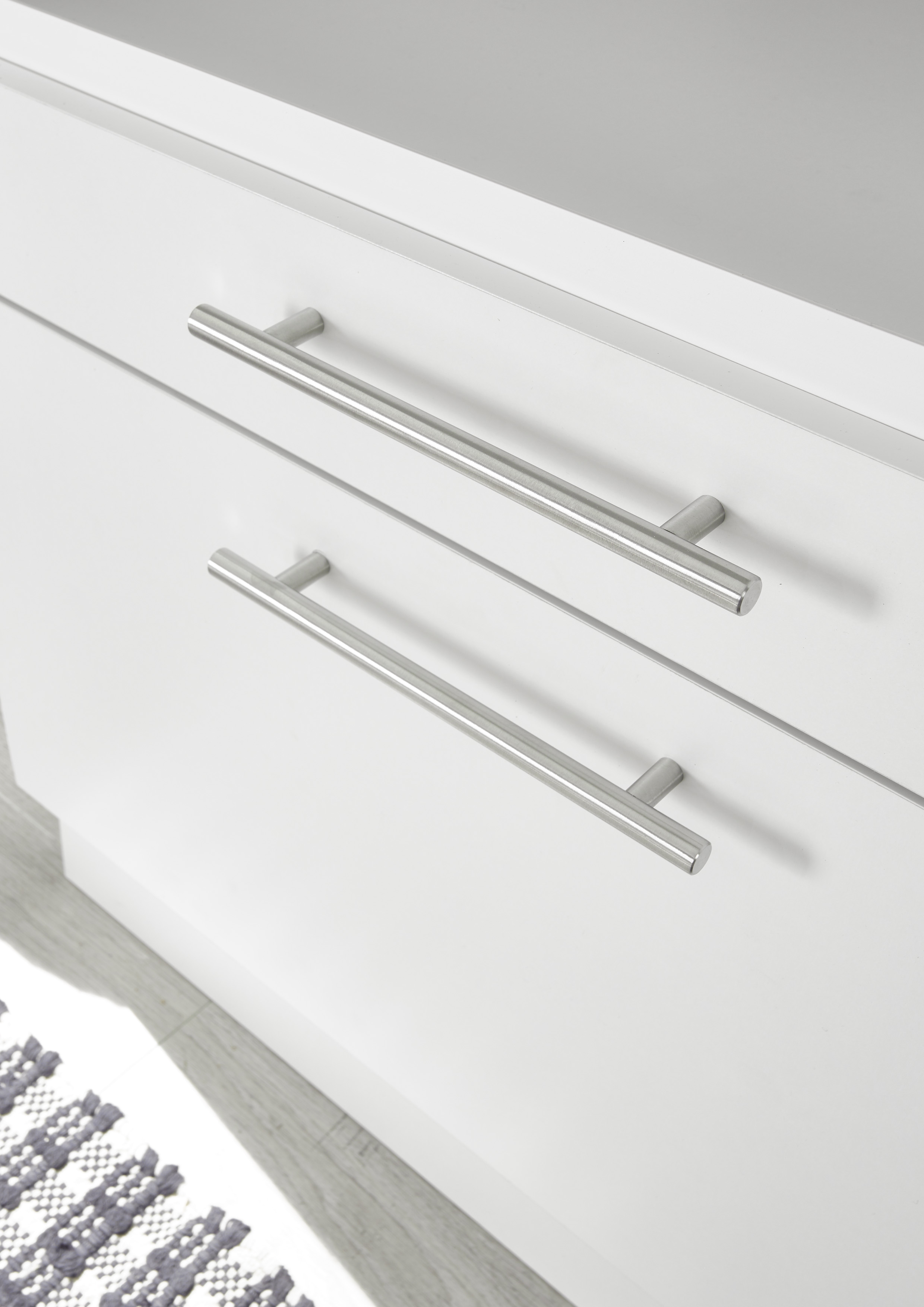 Weißer Schrank mit zwei silbernen Griffen. Der Schrank steht auf einem hellen Holzboden mit einem Teppich.