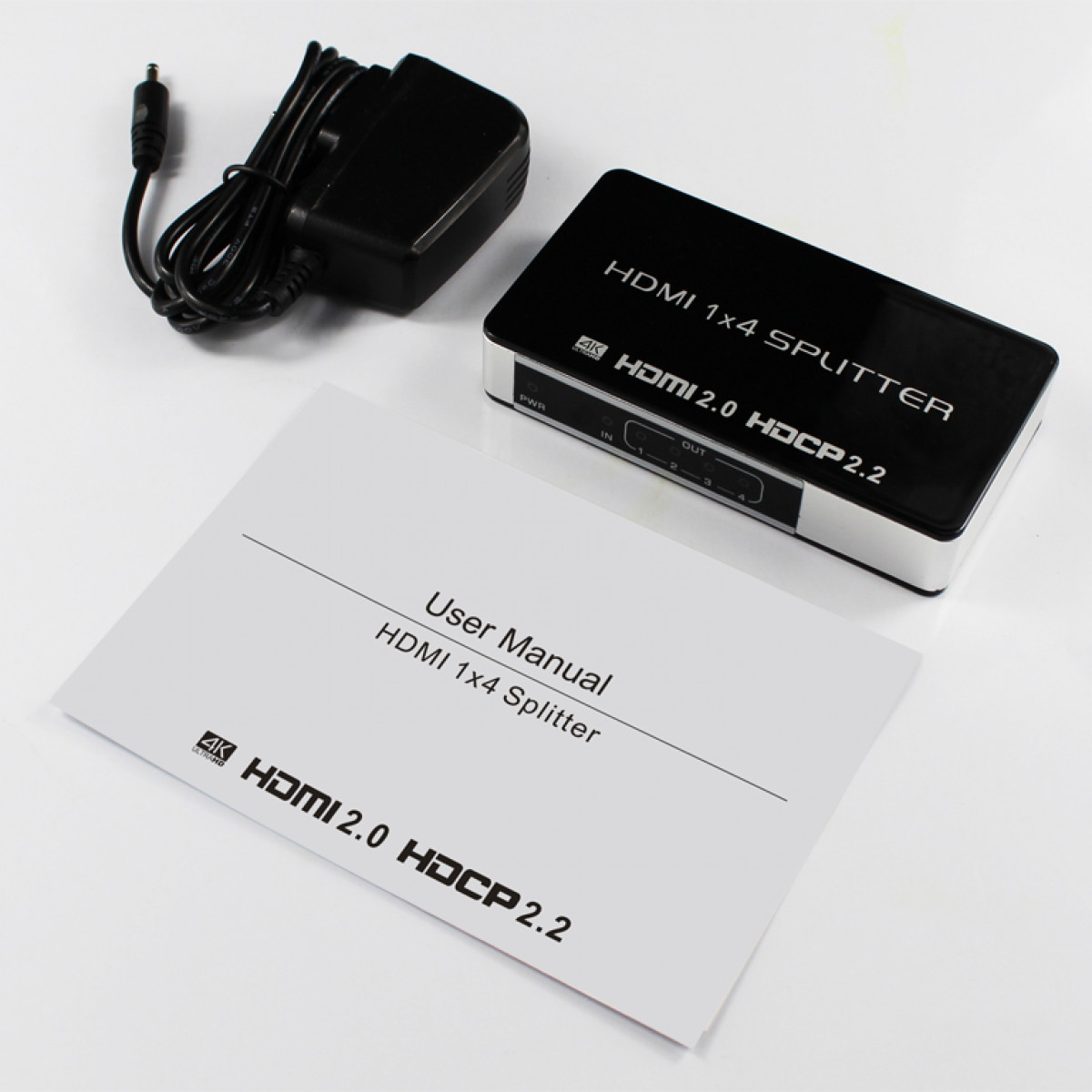 HDMI splitter with power adapter and a user manual on a white surface. Black and white.
