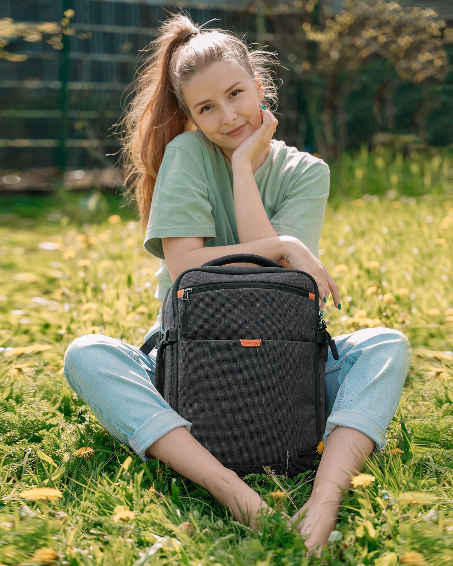 Eine Frau sitzt auf Gras mit einem Rucksack. Sie schaut in die Kamera, lächelt und trägt Freizeitkleidung.