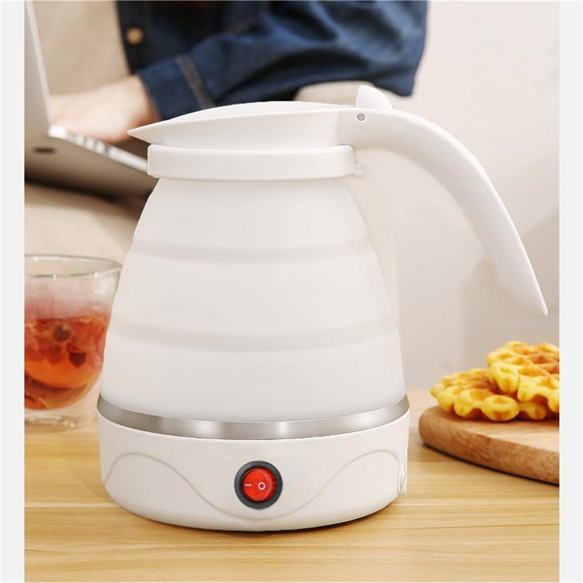 Hervidor eléctrico blanco y plegable. Un vaso de té y gofres en primer plano sobre una superficie de madera.