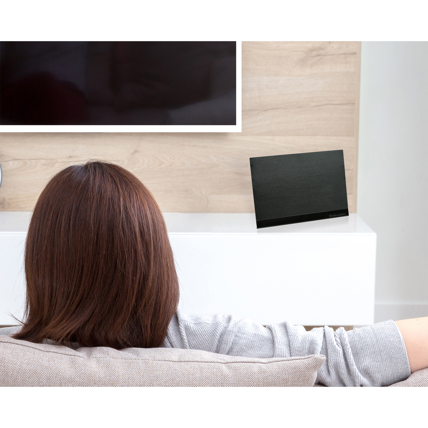 Kobieta siedzi na kanapie i ogląda telewizję. Na półce stoi czarny głośnik.