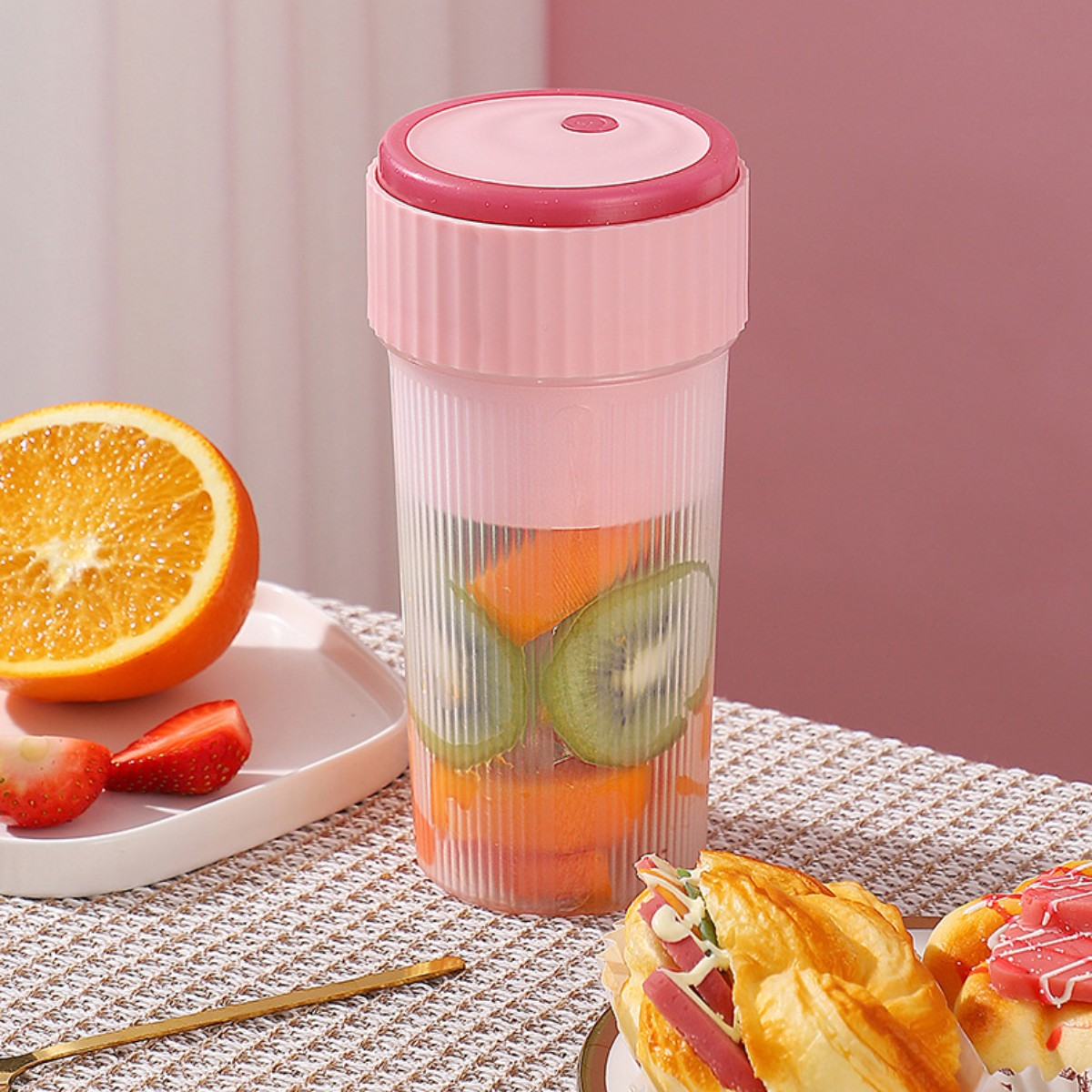 Licuadora rosa con frutas dentro y una mesa con otras frutas y pasteles.