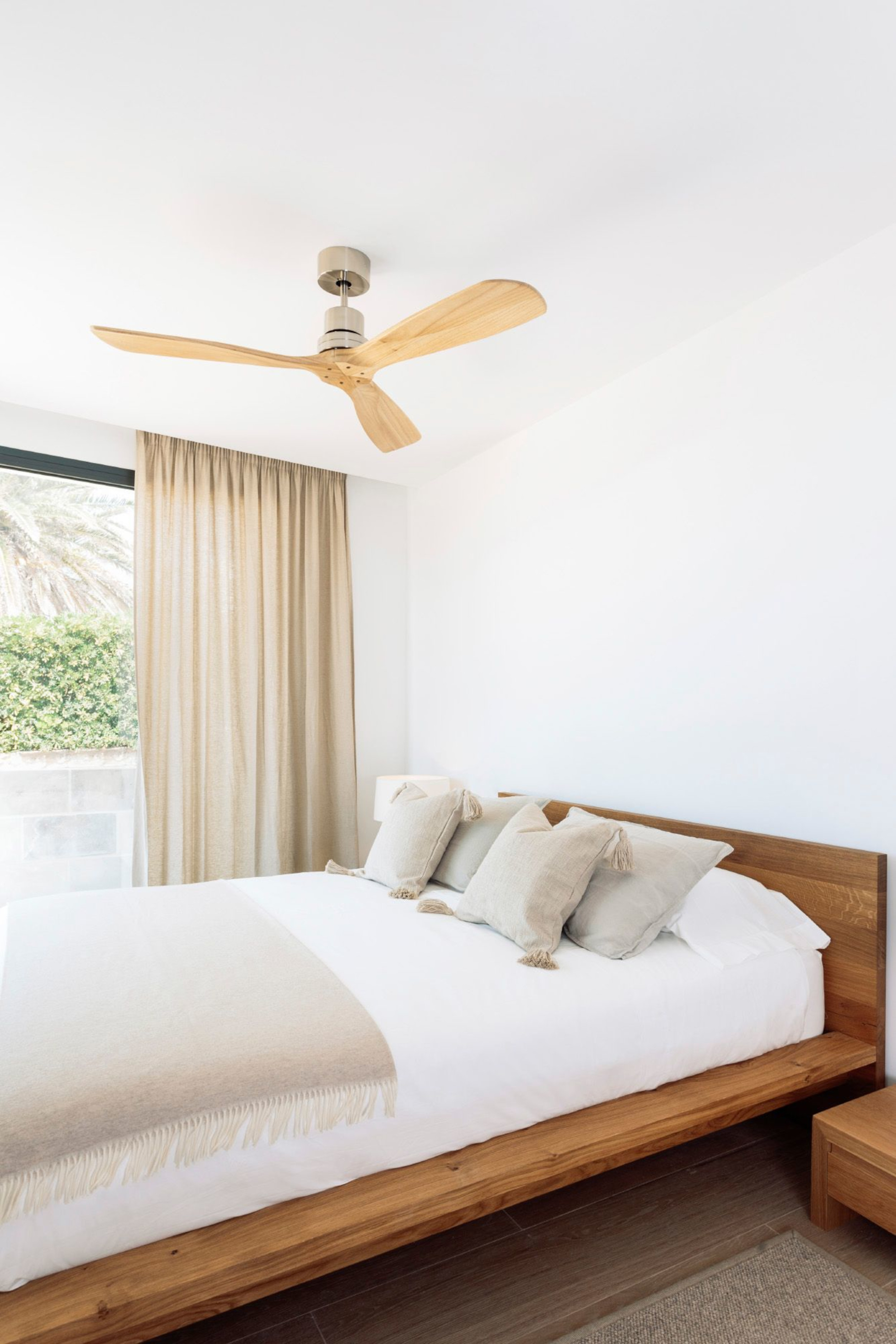 Una camera da letto con letto in legno, biancheria da letto bianca, tende beige e ventilatore a soffitto.