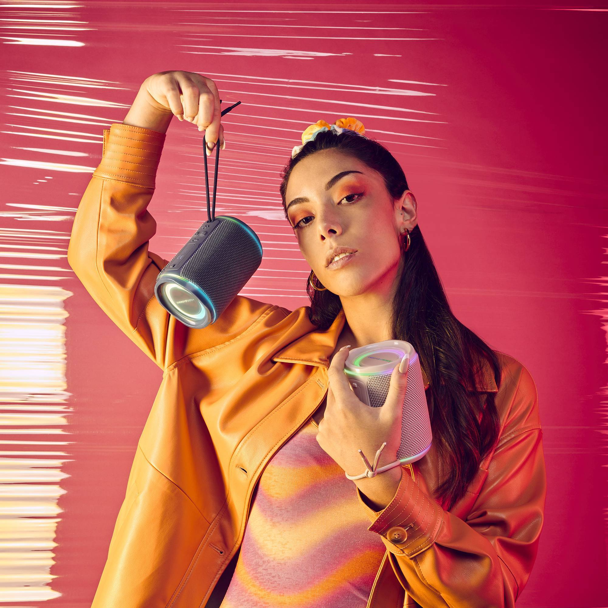 Kobieta w pomarańczowym trzymająca dwa cylindryczne głośniki z kolorowymi pierścieniami świetlnymi, na czerwonym tle.