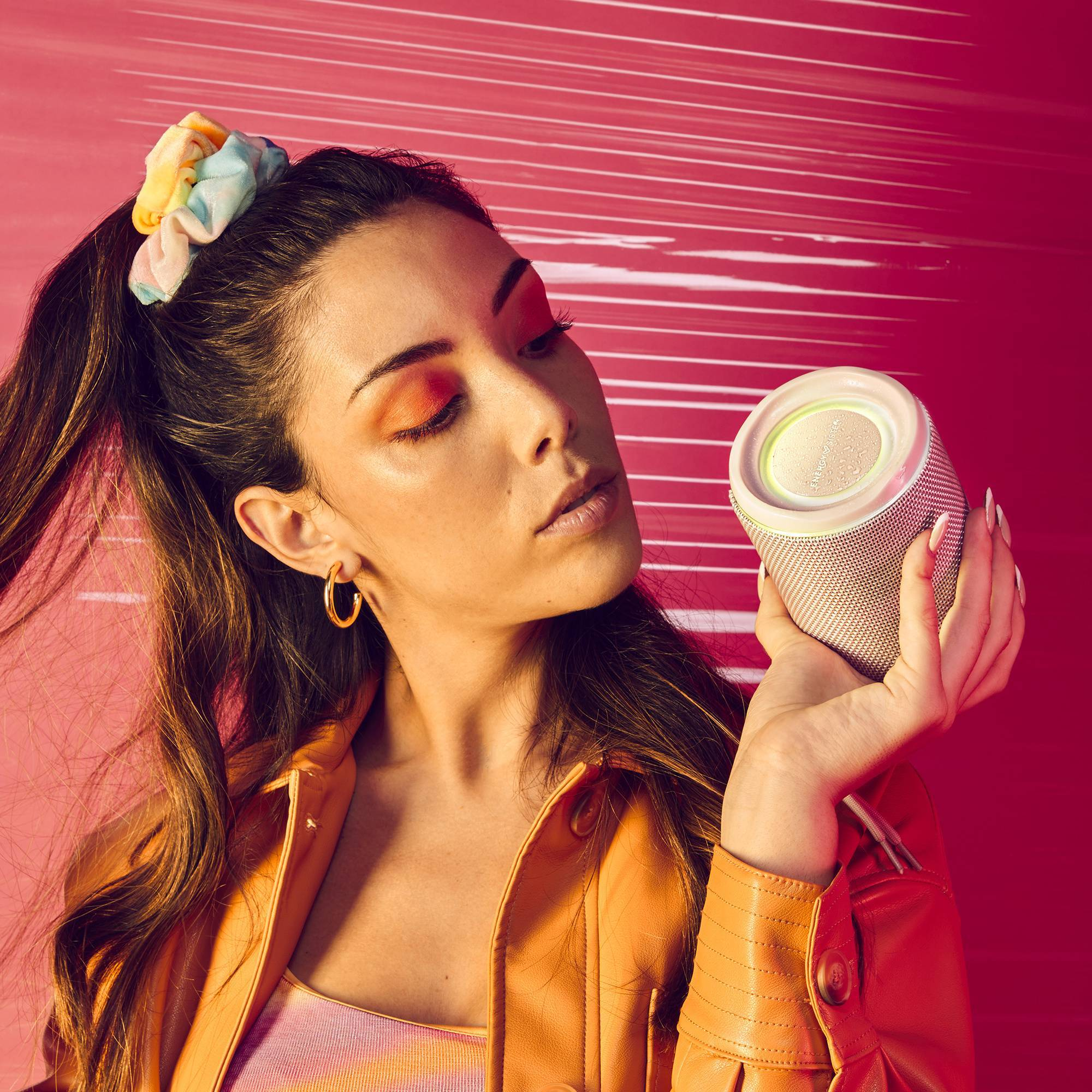 Kobieta trzymająca mały głośnik na różowym tle, z żółtą gumką do włosów.