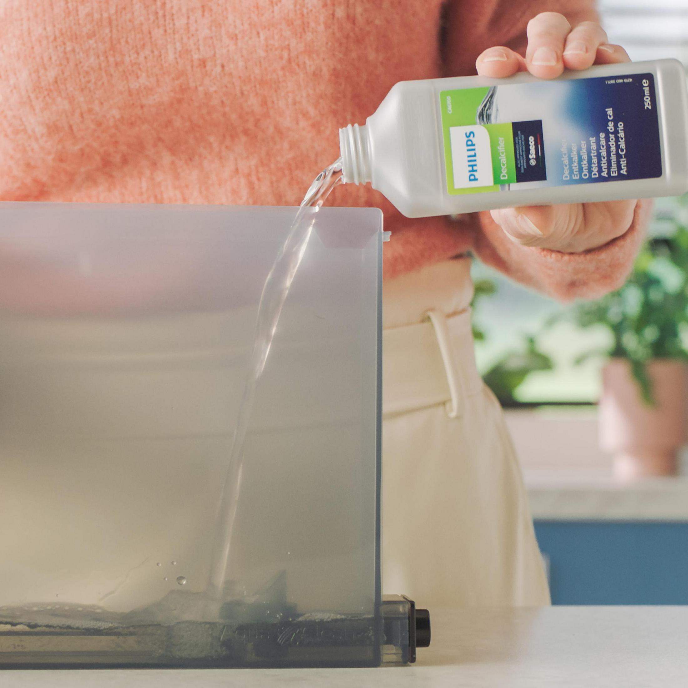 Eine Person gießt eine Entkalkerflüssigkeit aus einer weißen Flasche in einen Wassertank. Ein Etikett auf der Flasche ist sichtbar.