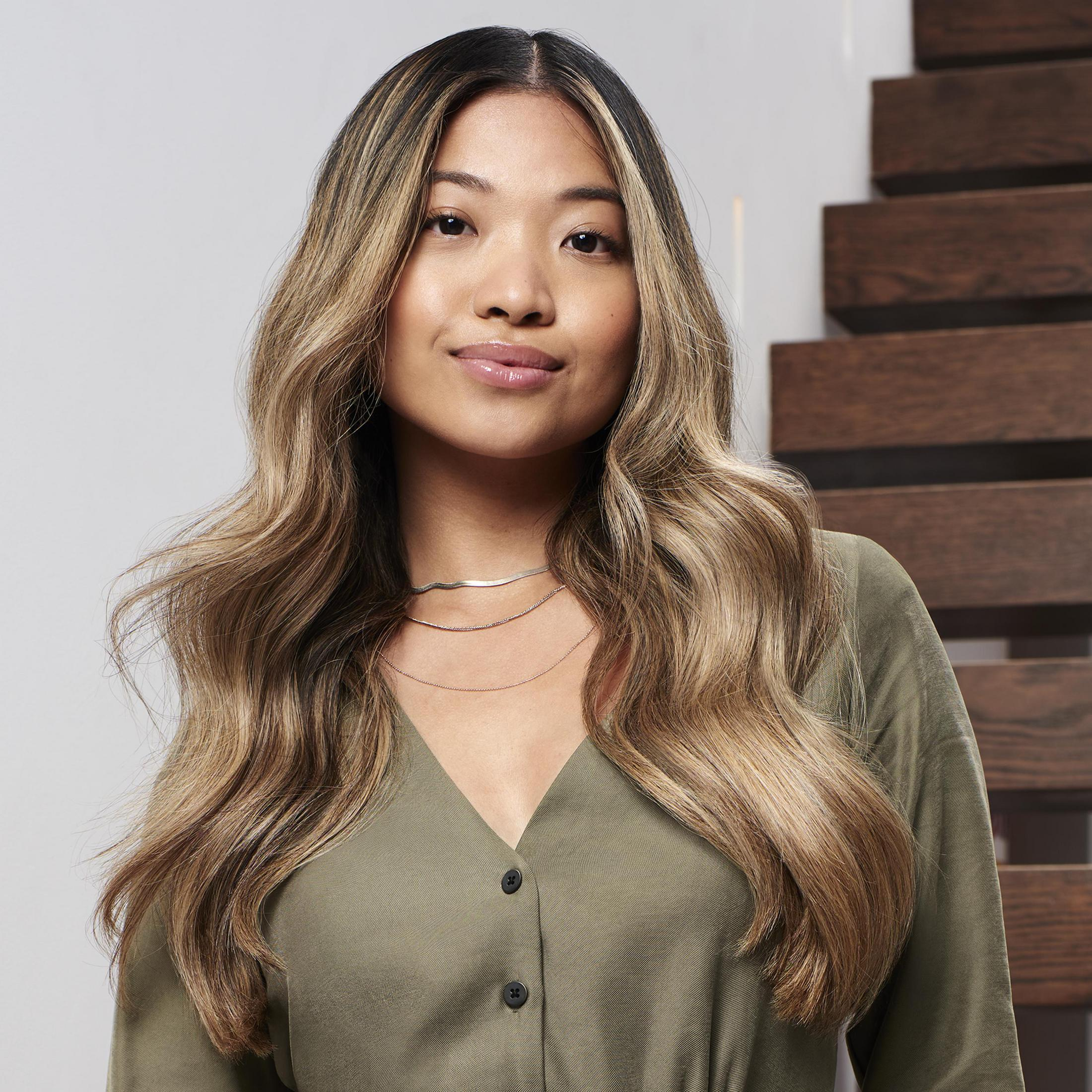 Młoda Azjatka z długimi, falowanymi włosami, w oliwkowej sukience, pozująca przed schodami.