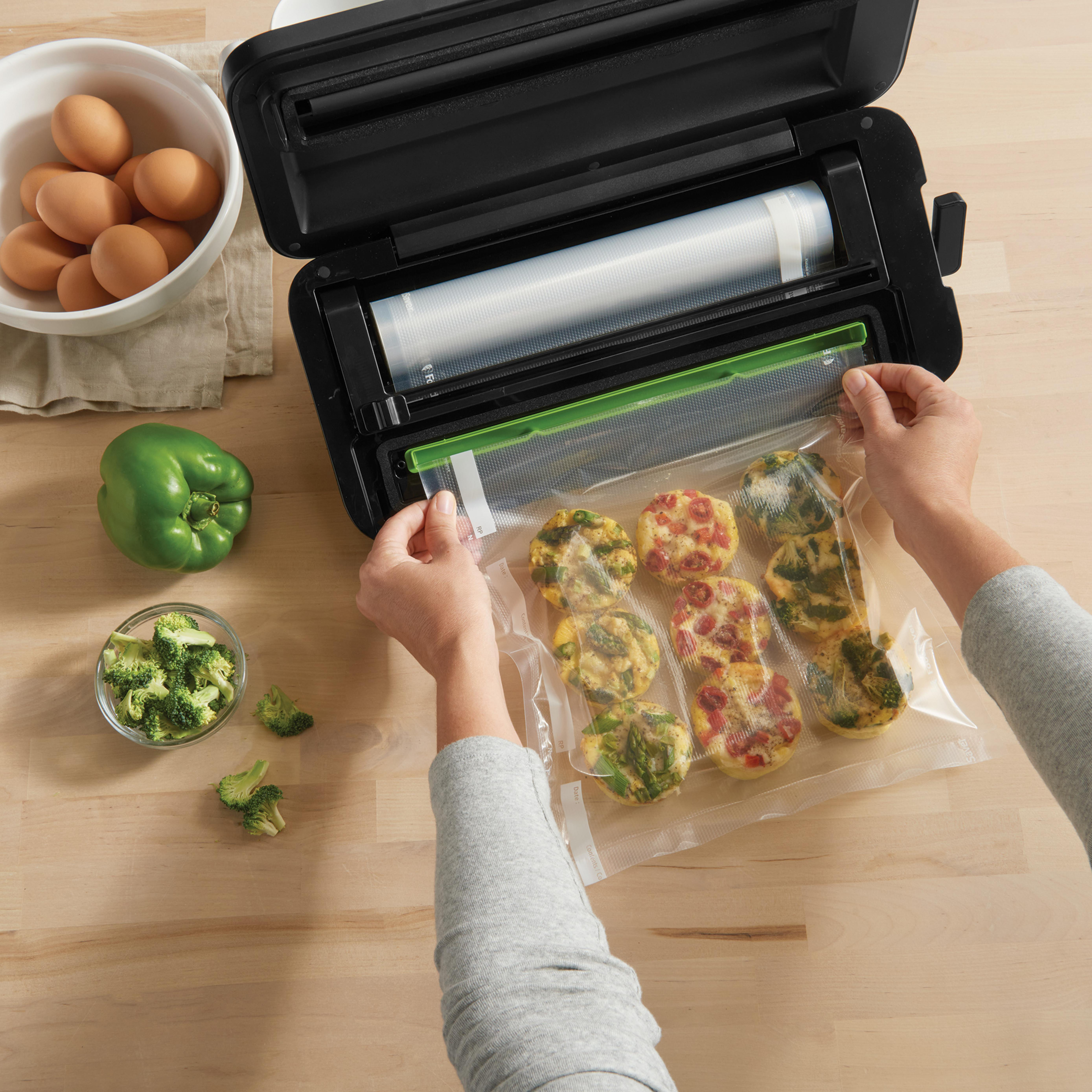 Hände halten eine Tüte mit Essen, neben Gemüse, Eiern und einem Vakuumierer auf einem Tisch.