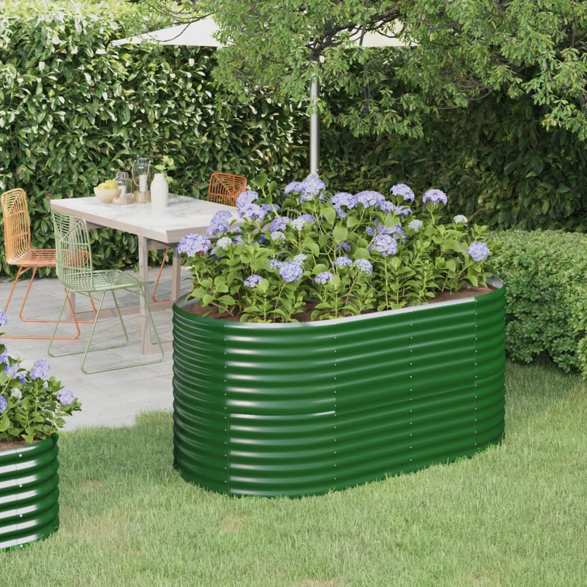 Grüner Metallpflanzer mit lila Blumen, Tisch im Freien, Stühle und grüner Heckenhintergrund.