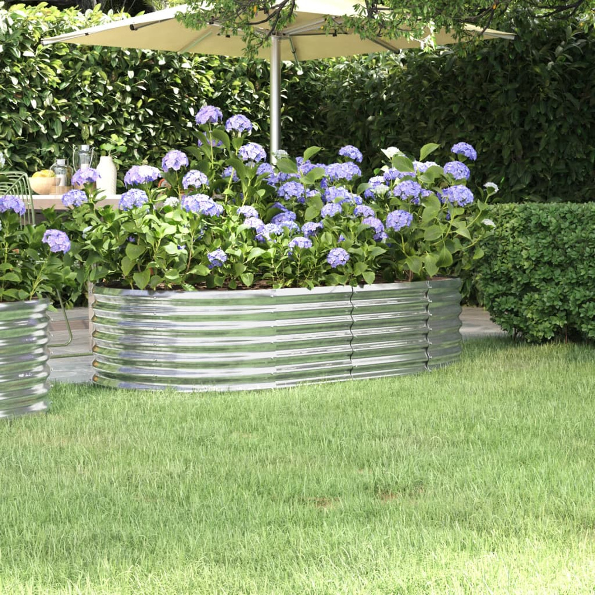 Metall-Pflanzkasten mit blauen Blumen in einem Garten unter einem Sonnenschirm. Grünes Gras und Hecke im Hintergrund.