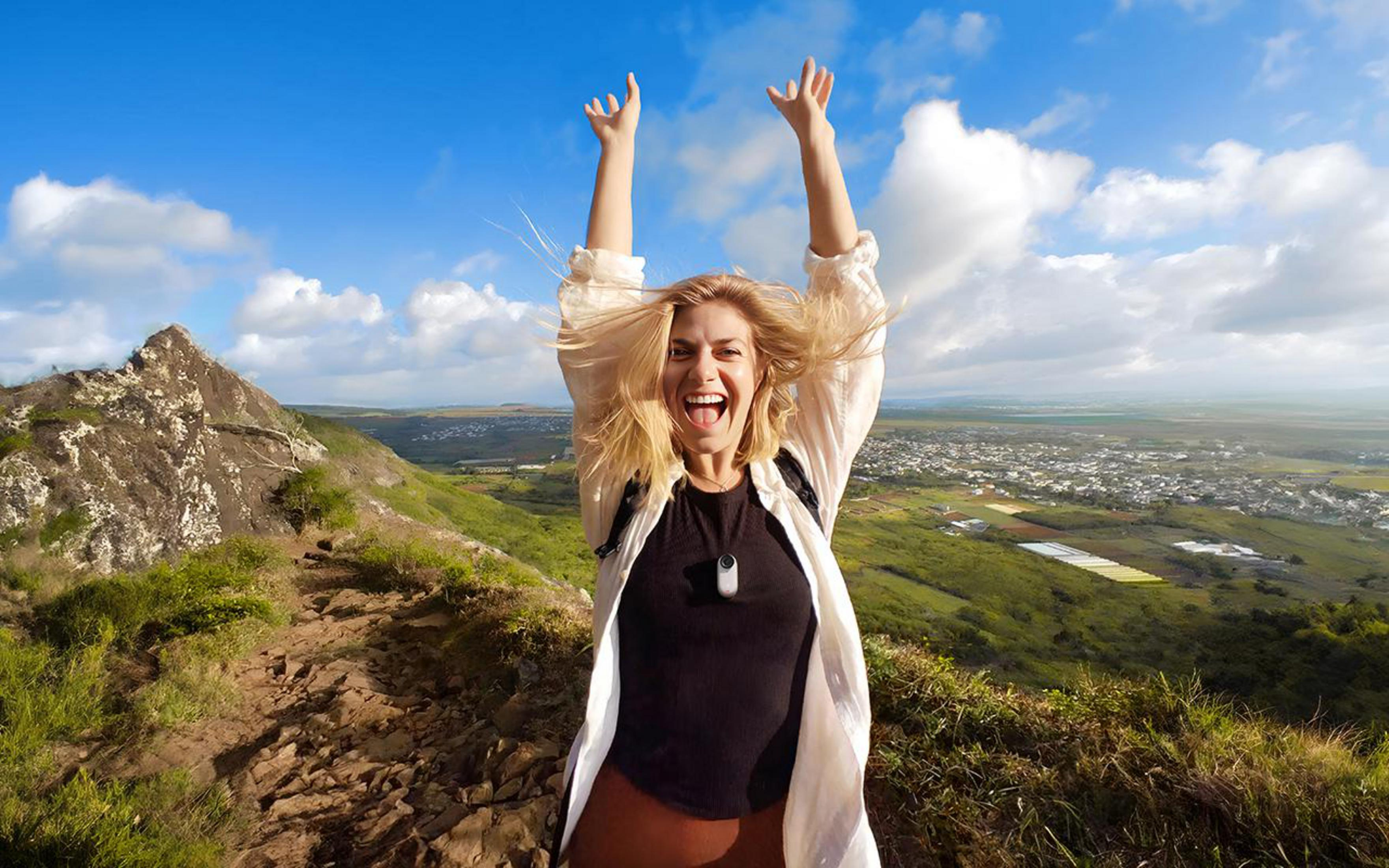 Kobieta z podniesionymi ramionami na górze, uśmiechnięta, z malowniczym krajobrazem wzgórz i nieba.