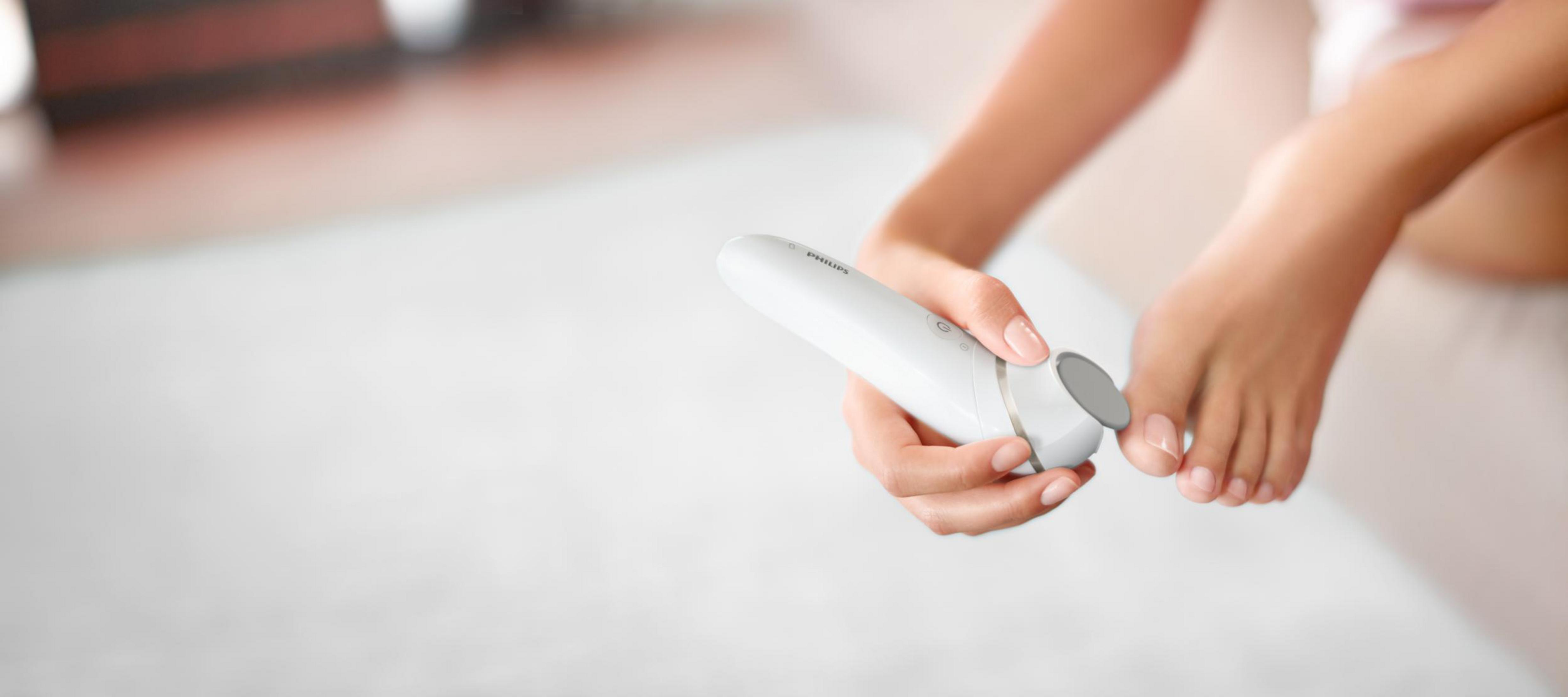 Osoba używająca białego pilnika do stóp, aby wygładzić stopę. Skupienie jest na stopie i urządzeniu.