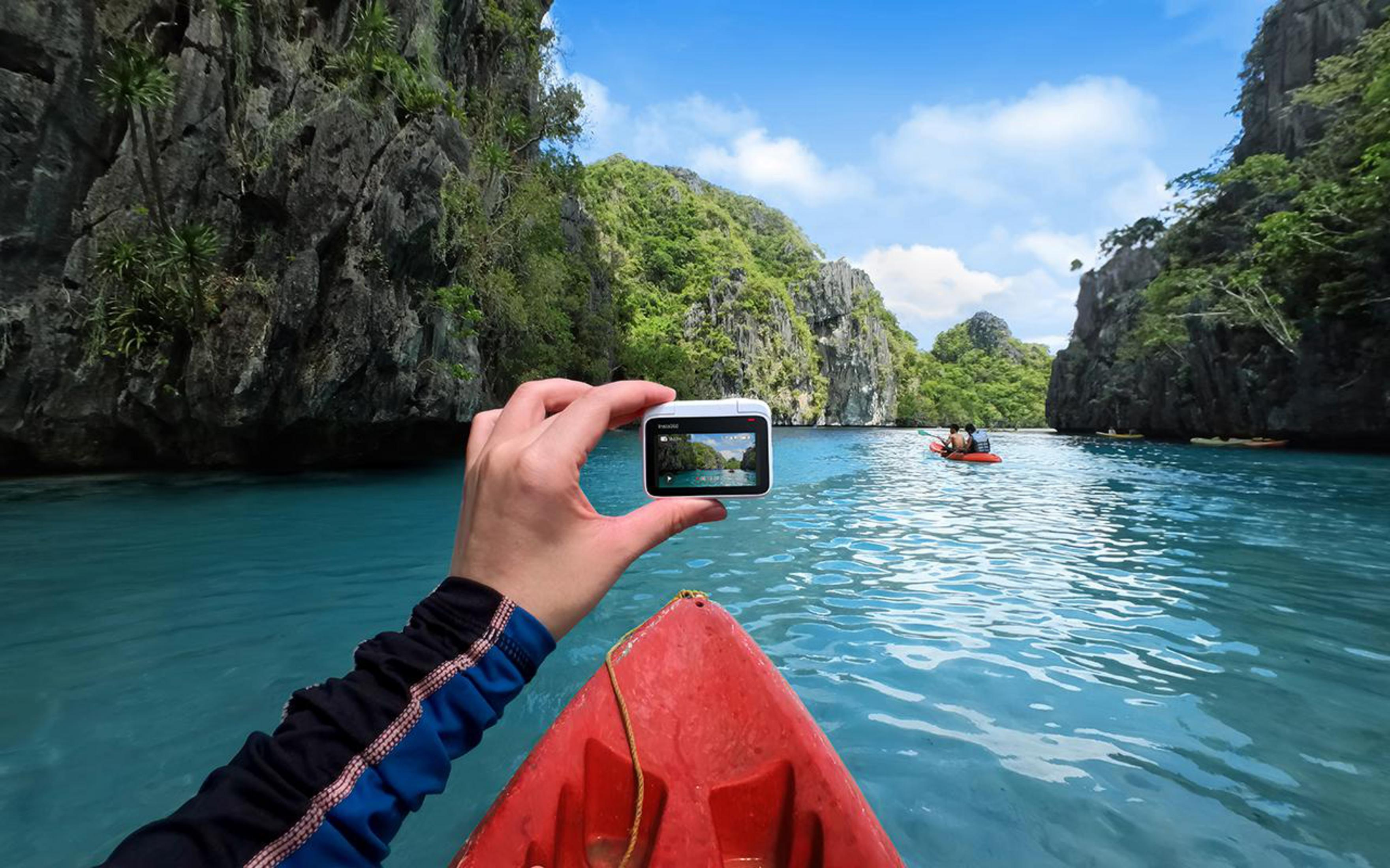 Hand holding camera, capturing turquoise water and lush islands. Red kayak in foreground, other boats in the distance.