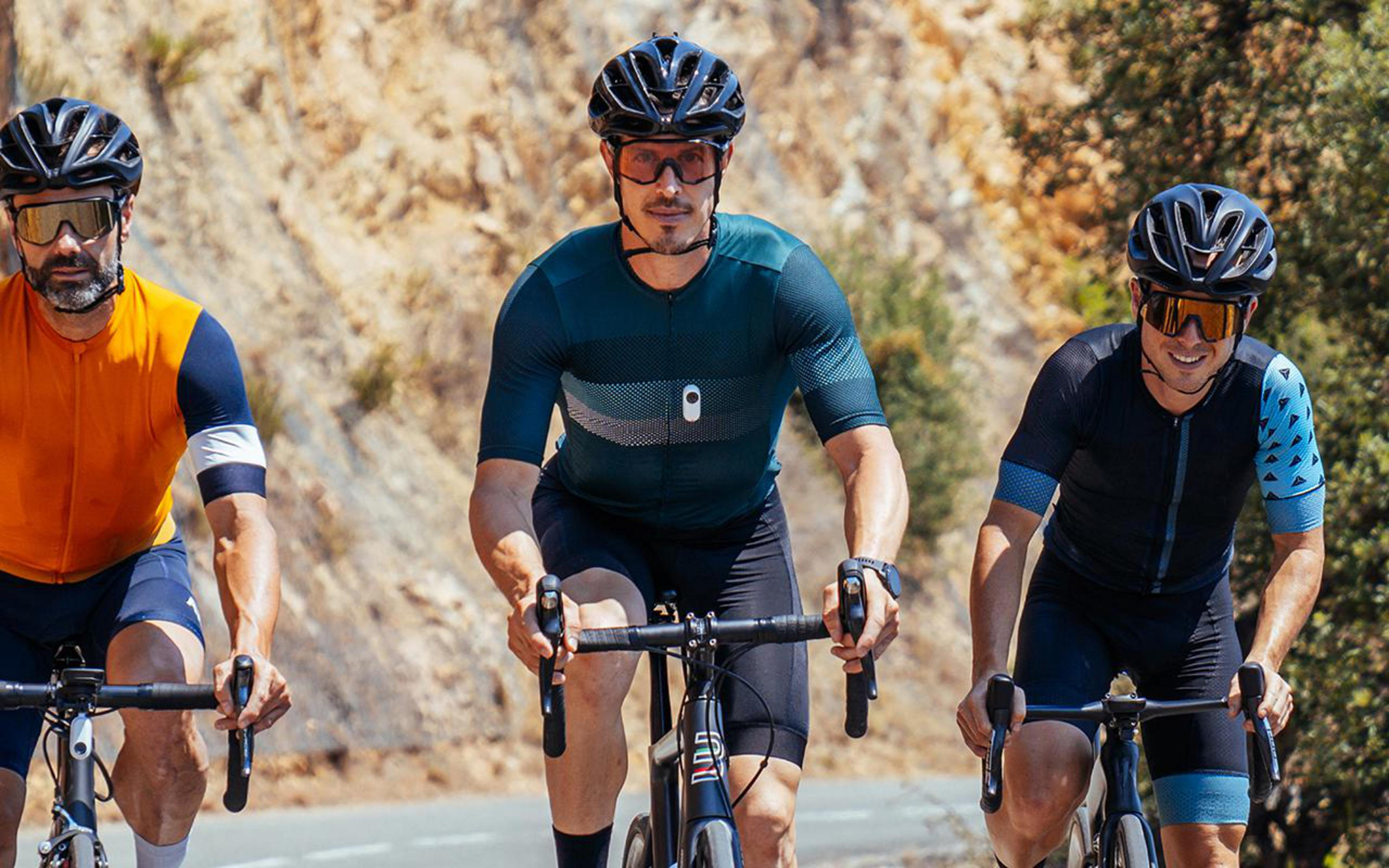 Three cyclists in cycling gear ride on a road. They are wearing helmets and sunglasses.
