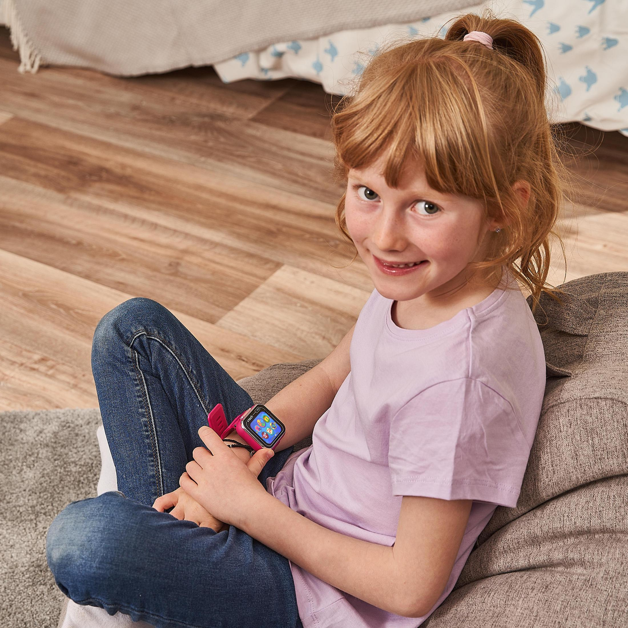Una bambina dai capelli rossi è seduta su un cuscino, indossa uno smartwatch rosa.
